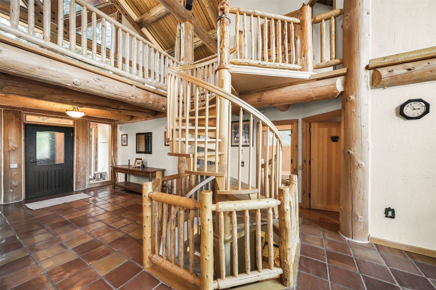 This rustic entryway features a prominent spiral staircase crafted from light wood logs, serving as the central architectural focal point. The space is finished with warm-toned square floor tiles and exposed log beams overhead, creating a cozy, cabin-like atmosphere. A lofted walkway with matching log railings overlooks the foyer, while a dark-framed front door and a wall-mounted clock add functional character to the open-concept layout.