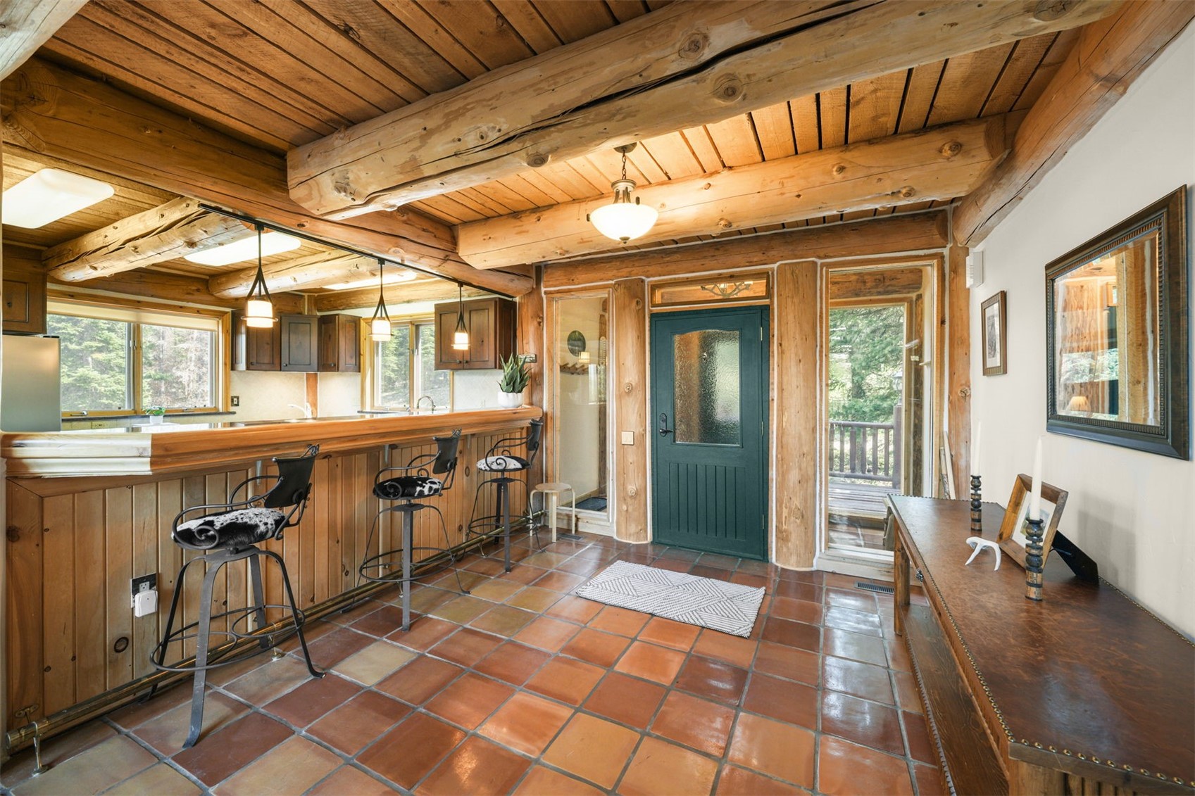 This rustic entryway features a warm, log-cabin aesthetic with exposed timber ceiling beams and terracotta-style floor tiles. A dark green front door serves as the focal point, flanked by glass panels that allow natural light to filter in, while a wooden bar counter with three stools creates a cozy transition space. The room is accented by a long wooden console table and a framed mirror, contributing to a welcoming and authentic mountain-lodge atmosphere.