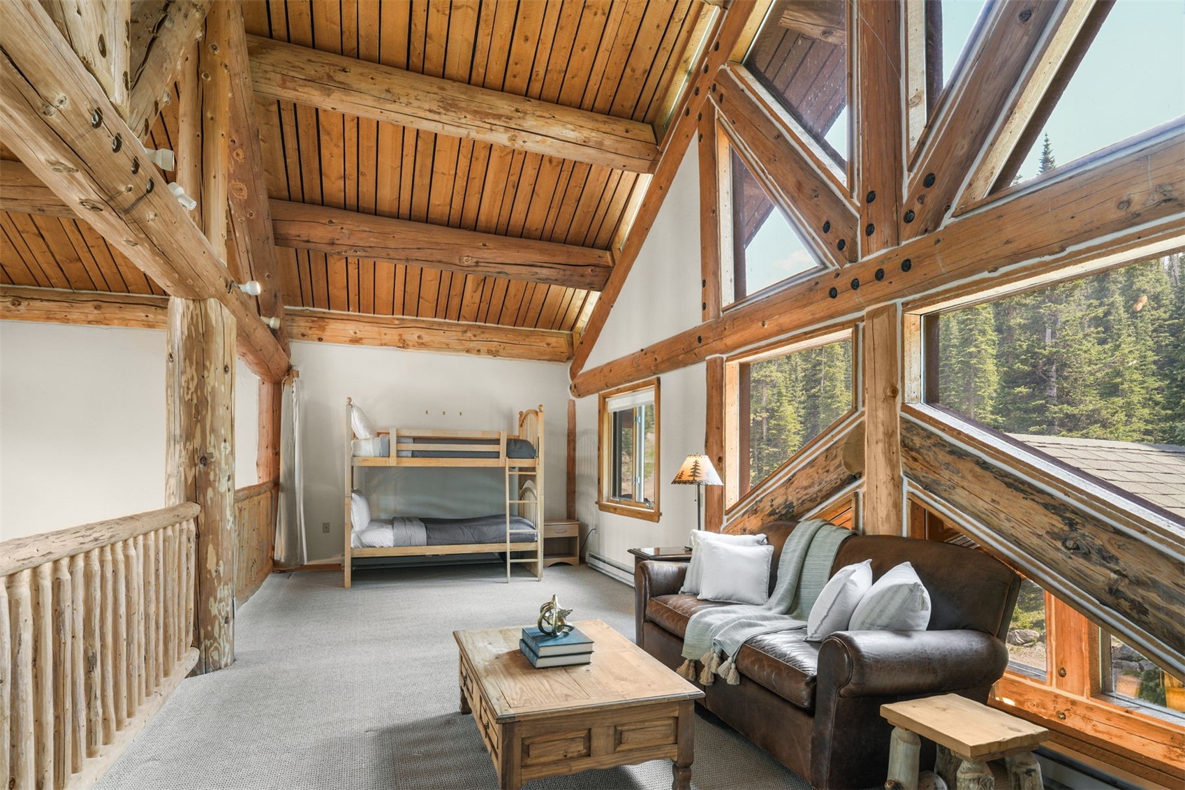 This rustic loft space features a cozy bunk bed and a comfortable leather sofa, creating a versatile living and sleeping area. The room is defined by its impressive timber-frame construction, exposed log beams, and a dramatic wall of large windows that frame the surrounding forest. The combination of light-toned wood ceilings, neutral carpeting, and natural light gives the space a warm, inviting mountain-lodge atmosphere.