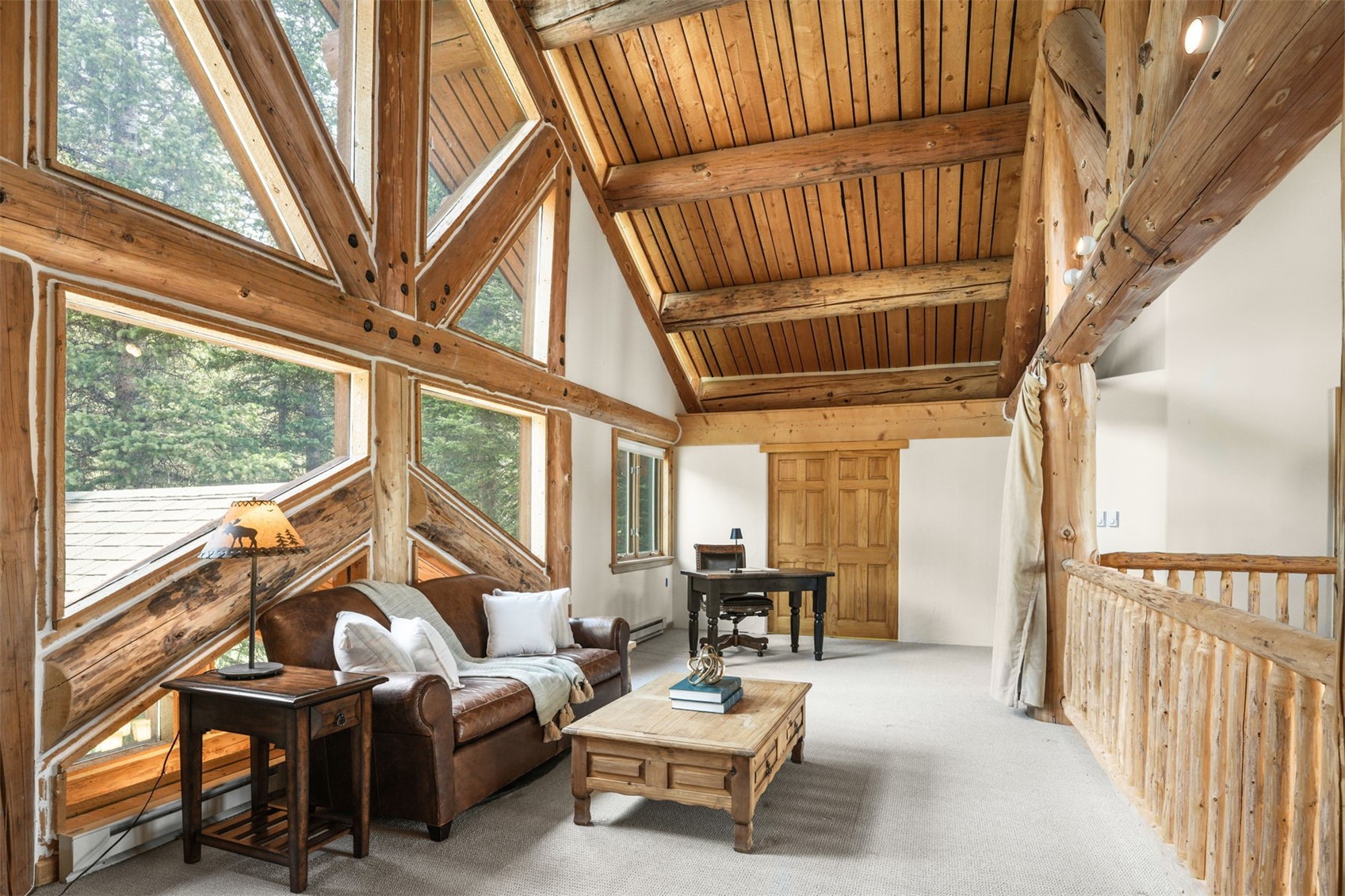 This rustic loft space features a vaulted ceiling with exposed timber beams and a warm, wood-paneled finish. A leather sofa and wooden coffee table create a cozy seating area, while a desk is positioned near the back wall for a functional workspace. Large, angular windows provide expansive views of the surrounding forest, filling the room with natural light.