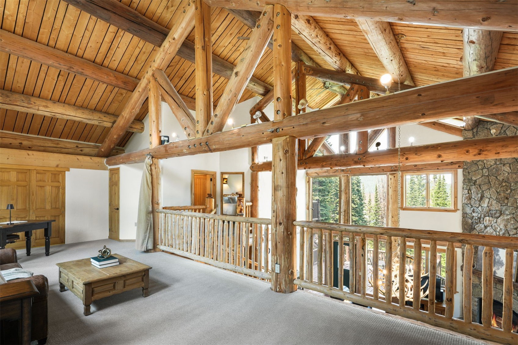 This rustic loft space features a vaulted ceiling with exposed log beams and a light wood plank ceiling. A wooden railing overlooks the lower level, while a stone-clad chimney breast adds texture to the room. The area is furnished with a coffee table and a desk, creating a cozy, open-concept landing area with views through large windows.