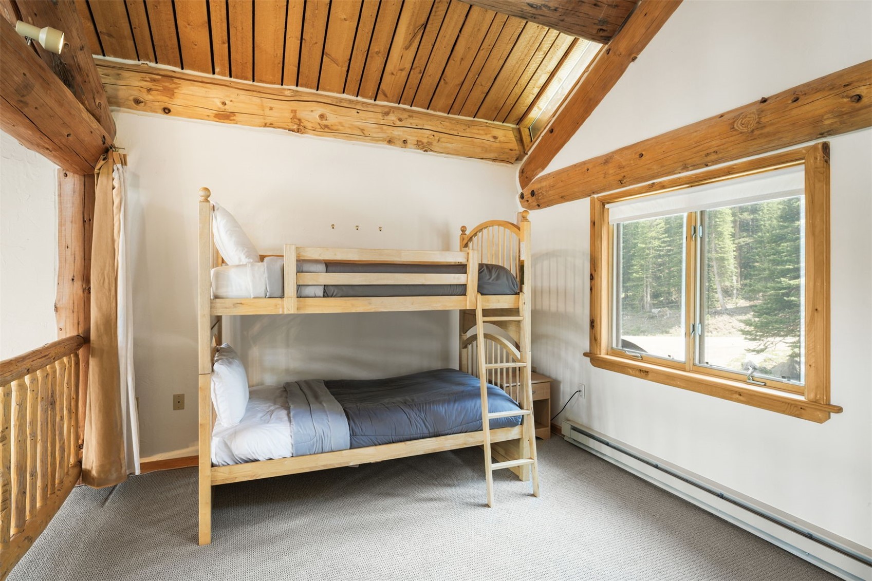 This cozy bedroom features a light wood bunk bed set as the central focus, positioned against a white wall under a vaulted ceiling with exposed wooden beams. A large window to the right provides natural light and a view of the surrounding forest, while the room is finished with neutral carpeting and a rustic wooden railing on the left. The space offers a warm, cabin-like atmosphere suitable for a guest room or children's sleeping area.
