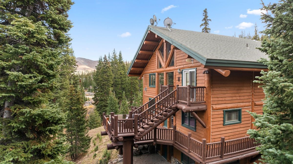 This side view of a rustic log cabin showcases its multi-level design nestled within a dense forest of evergreen trees. The structure features a prominent wooden staircase leading to a mid-level deck, with a smaller balcony above and a stone-foundation base below. The setting is mountainous and serene, emphasizing the home's integration into the natural landscape.