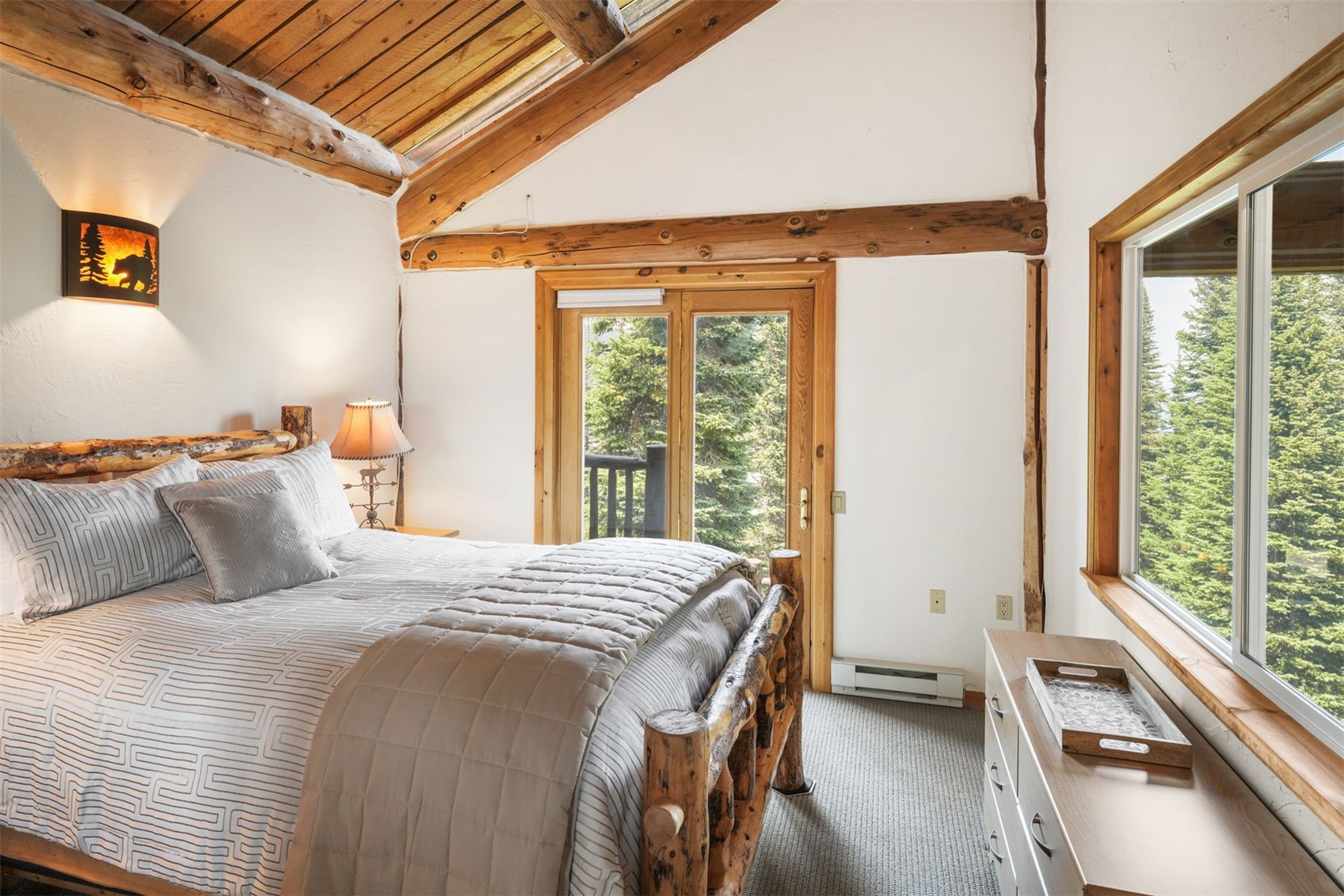 This rustic bedroom features a log-frame bed with a patterned quilt and a matching wooden dresser. The room is highlighted by exposed wooden ceiling beams, a vaulted ceiling, and a glass door that opens to a balcony with forest views. A large window on the right provides ample natural light, creating a cozy and serene atmosphere.