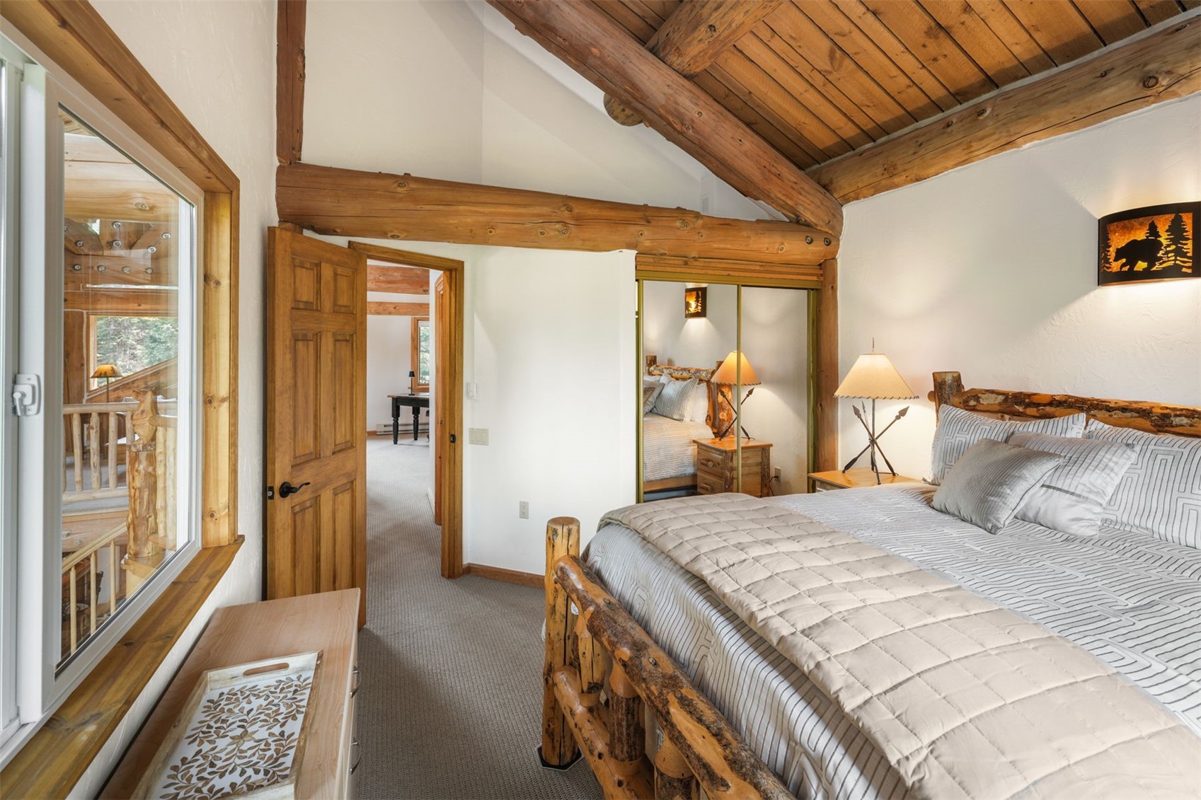 This rustic-style bedroom features a log-frame bed and exposed timber ceiling beams, creating a cozy, cabin-like atmosphere. The room includes a mirrored closet, a bedside lamp, and a window looking out onto an interior balcony area, while a doorway leads to an adjacent room. The neutral color palette and natural wood textures provide a warm and inviting aesthetic.