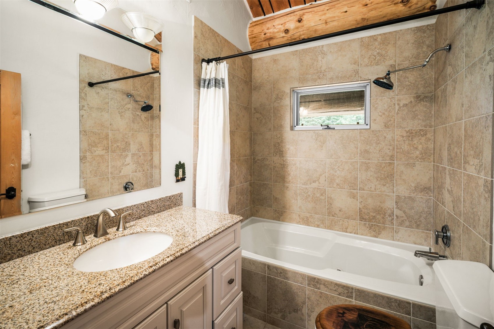This bathroom features a vanity with a stone countertop and a white undermount sink, paired with a mirror reflecting the shower area. The tub-shower combination is enclosed by a white curtain and surrounded by neutral-toned wall tiles, with a small window providing natural light. A rustic exposed wooden beam runs across the ceiling, adding a warm, cabin-like aesthetic to the space.
