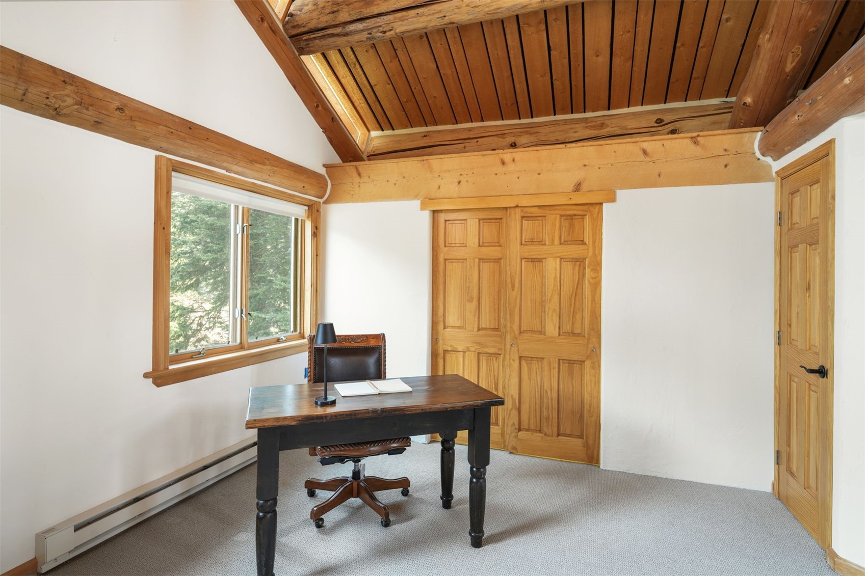 This home office features a rustic aesthetic with exposed wooden beams and a vaulted wood-paneled ceiling. The room is furnished with a dark wood desk and a leather office chair, positioned near a window that provides natural light and a view of the outdoors. The space is finished with white walls and neutral carpeting, creating a quiet and focused atmosphere.