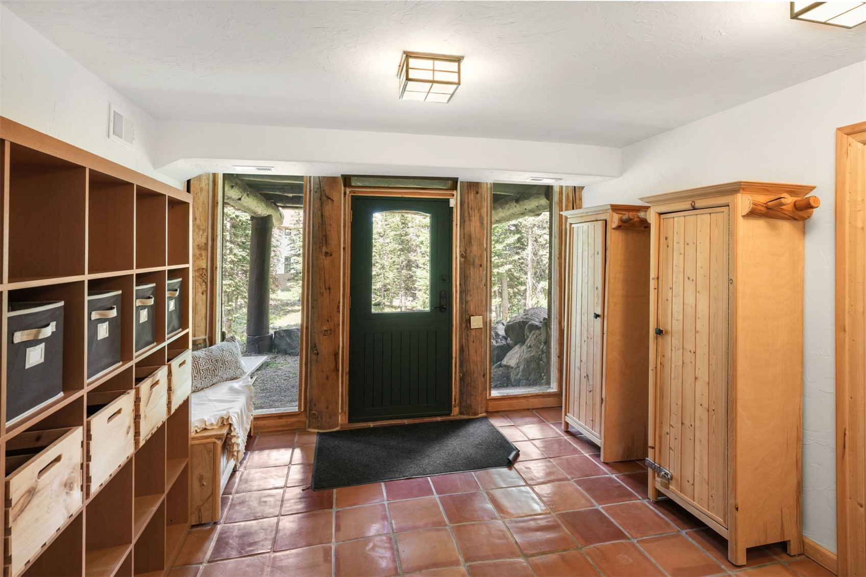 This rustic entryway features terracotta-style tile flooring and a dark green front door flanked by tall windows that provide a view of the surrounding forest. The space is furnished with a built-in storage unit with cubbies, a wooden bench with a patterned cushion, and two tall wooden cabinets for additional organization. The warm wood tones and natural light create a welcoming, cabin-inspired atmosphere.