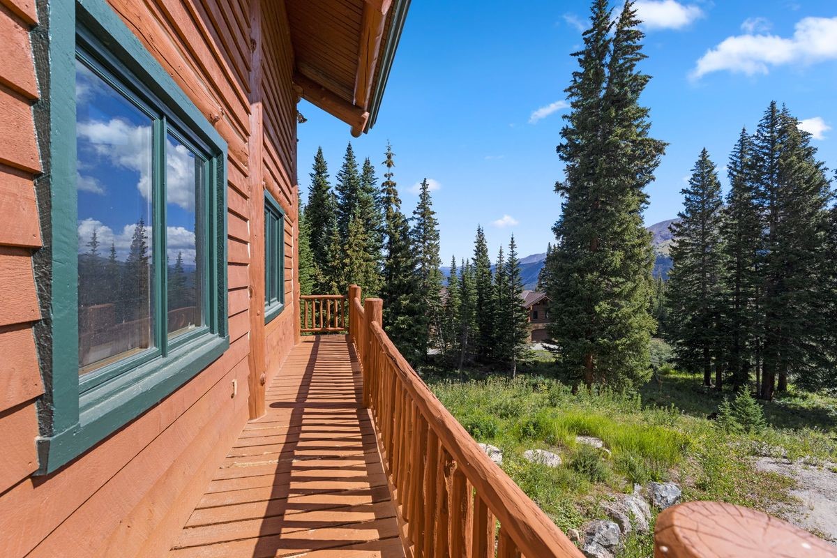 This image captures a rustic wooden balcony attached to a log-style home, offering a panoramic view of a lush, forested mountain landscape. The deck features a sturdy log railing and provides a clear vantage point of the surrounding pine trees and distant peaks under a bright blue sky. The perspective is from the balcony looking outward, emphasizing the peaceful, secluded nature of the property.