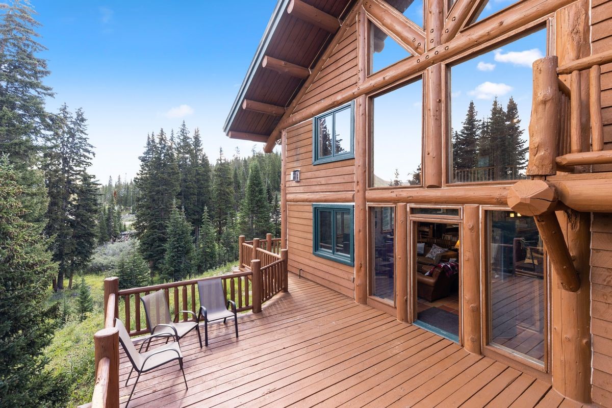 This image captures a spacious wooden deck attached to a rustic log cabin, offering a serene view of a dense pine forest. The deck features two metal-framed chairs and a wooden railing, while the cabin itself showcases large windows and a prominent log-beam construction. The perspective is from the deck looking toward the side of the house, highlighting the seamless integration between the home's architecture and the surrounding natural landscape.