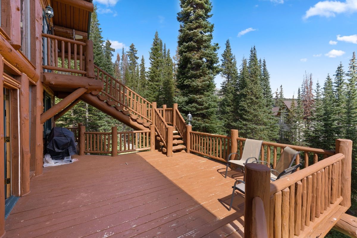 This spacious, multi-level wooden deck features rustic log railings and stairs leading to an upper balcony. The deck is furnished with two chairs and includes a covered grill, offering a scenic view of the surrounding pine forest and clear blue sky. The warm-toned wood construction creates a cozy, mountain-lodge aesthetic perfect for outdoor relaxation.