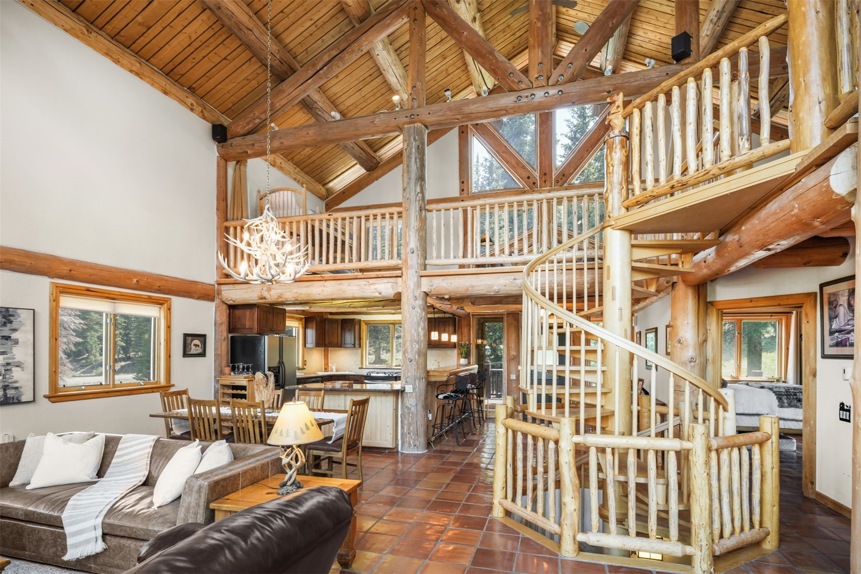 This expansive, rustic-style living area features a soaring vaulted ceiling with exposed log beams and a prominent antler chandelier. The open-concept layout seamlessly connects the living space to a dining area and kitchen, all anchored by a central spiral staircase with log railings. Large windows and a lofted balcony enhance the airy, mountain-lodge atmosphere, while the terracotta-tiled floor adds warmth to the interior.