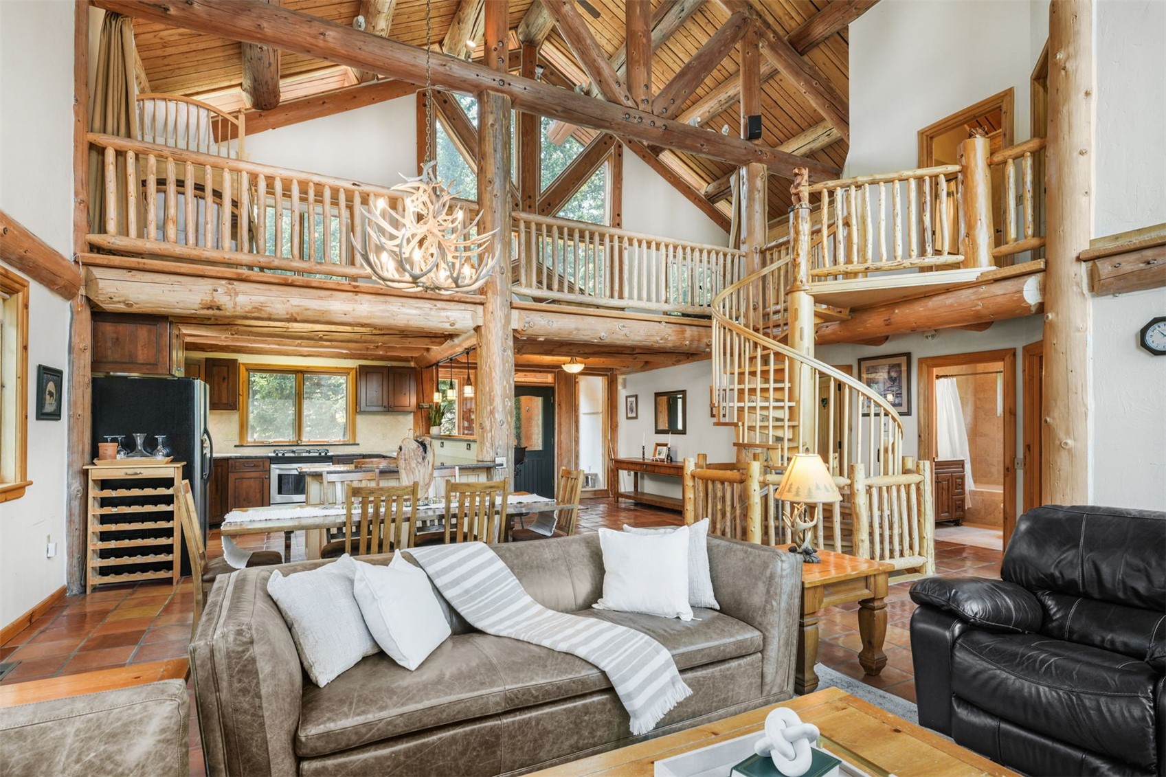 This expansive, open-concept living area showcases a rustic log cabin aesthetic with exposed timber beams and a vaulted ceiling. The space features a comfortable leather sofa, a spiral staircase leading to a lofted walkway, and an integrated kitchen and dining area in the background. Large windows and a distinctive antler chandelier enhance the lodge-like atmosphere, creating a warm and inviting mountain retreat feel.