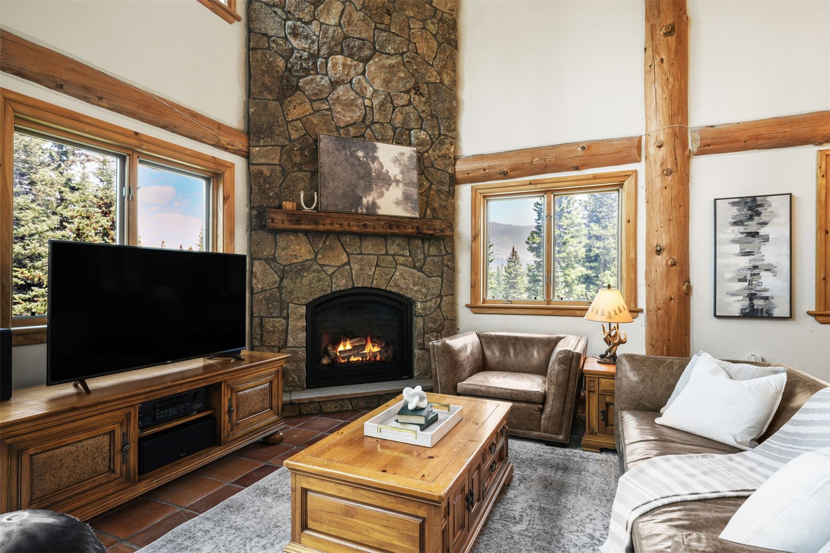 This cozy living room features a prominent stone-clad chimney breast that serves as the focal point, complemented by warm wood trim and exposed log beams. The space is furnished with a leather armchair, a matching sofa, and a sturdy wooden coffee table, all set upon a textured area rug. Large windows on either side of the fireplace provide ample natural light and views of the surrounding forest, creating a rustic and inviting atmosphere.