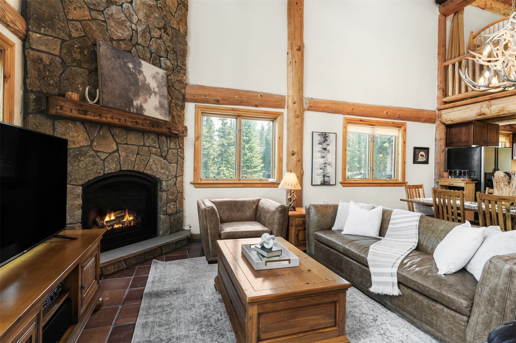 This cozy, rustic living room features a prominent stone-clad chimney breast with a wood mantel and a warm, glowing fireplace. The space is anchored by a large leather sofa and a matching armchair, arranged around a wooden coffee table on a textured area rug. Tall windows provide a view of the surrounding forest, while exposed timber beams and high ceilings enhance the mountain-lodge aesthetic.