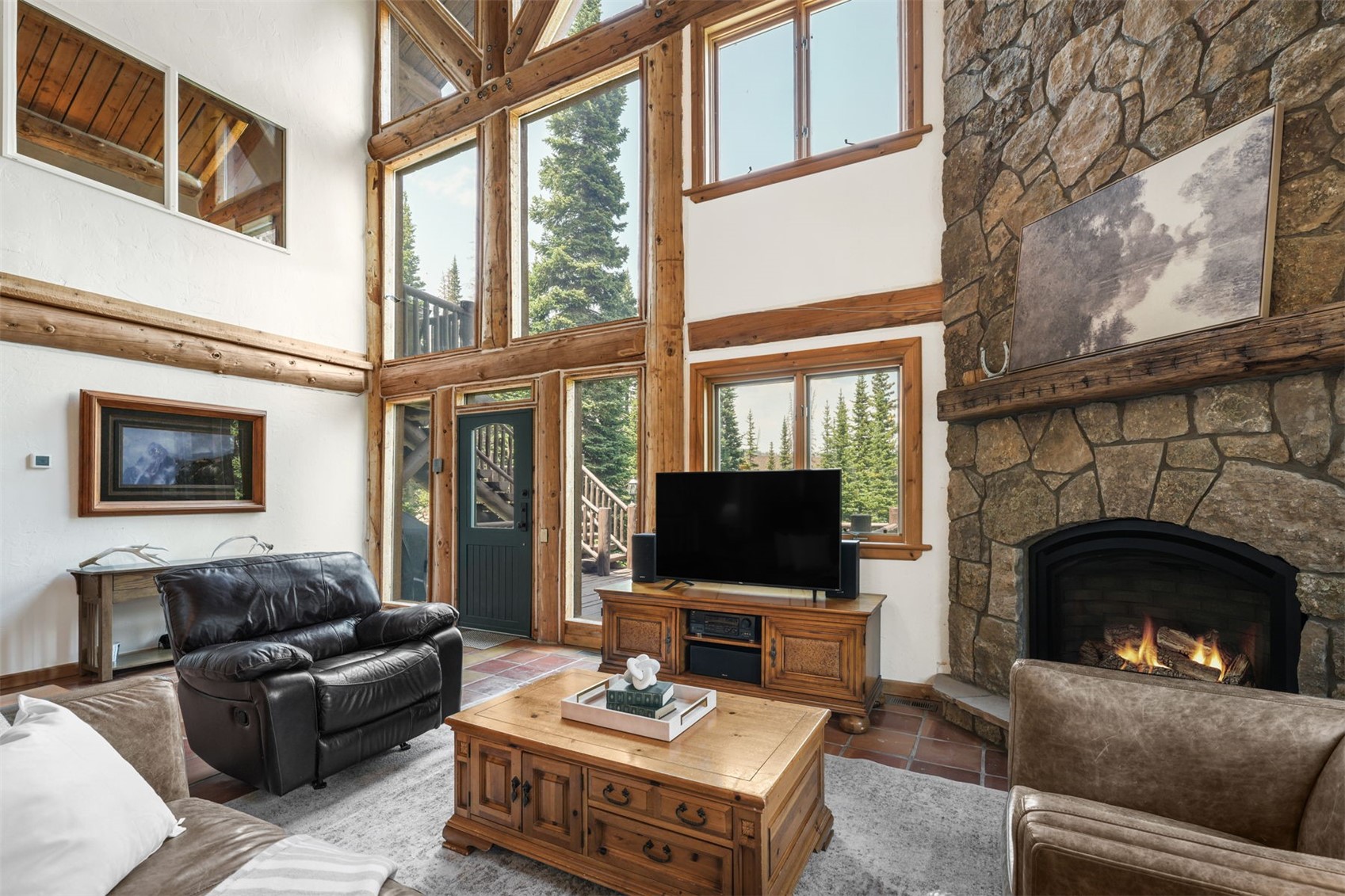 This spacious living room features a rustic aesthetic with exposed timber beams and a prominent stone-clad chimney breast housing a cozy fireplace. Large windows and a glass-paneled door allow for abundant natural light and views of the surrounding forest, while a leather armchair and a wooden coffee table anchor the seating area. The room is finished with tile flooring and a neutral area rug, creating a warm and inviting atmosphere.
