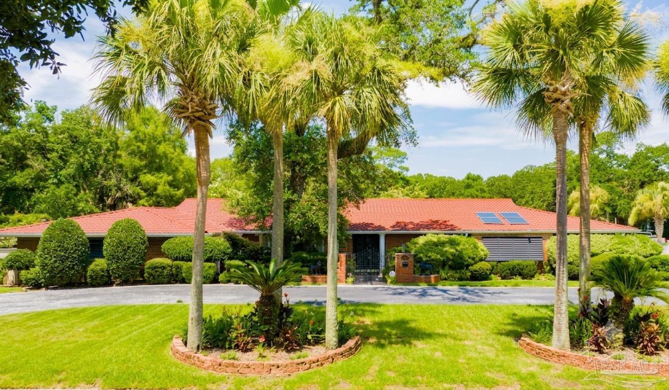 This front-facing view captures a sprawling, single-story ranch-style home with a distinctive red tile roof and brick exterior. The property is framed by mature palm trees and lush landscaping, with a paved driveway leading to a central gated entryway. The scene is bathed in bright daylight, highlighting the home's classic architectural style and well-maintained curb appeal.