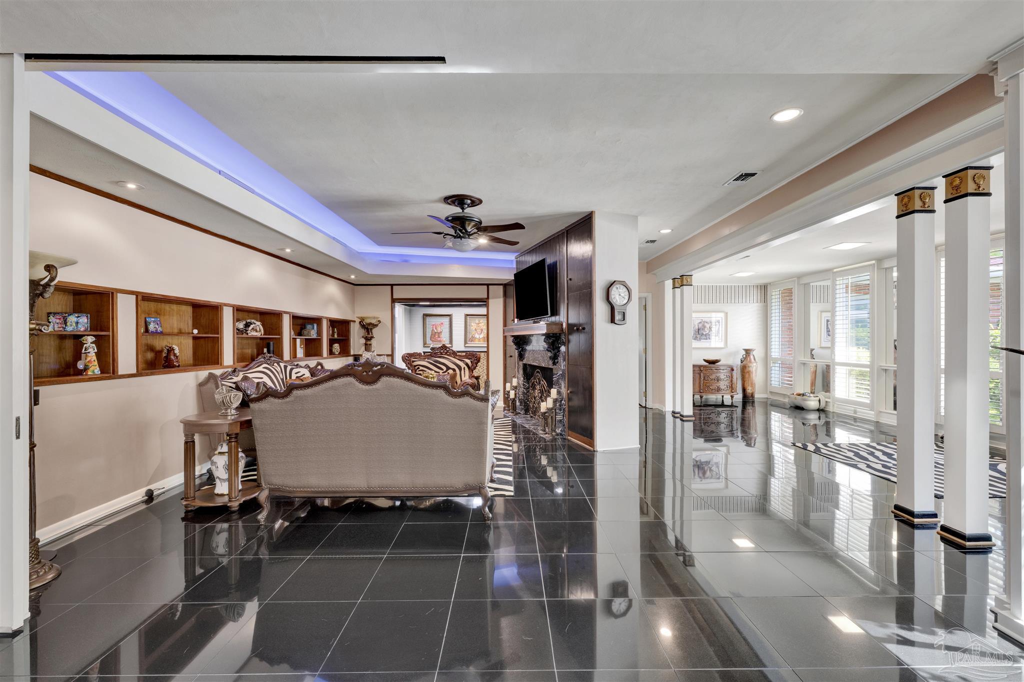 This spacious living area features high-gloss black tile flooring and a recessed ceiling with integrated blue accent lighting. The room is anchored by a traditional sofa with patterned upholstery, a dark wood fireplace surround with a mounted television, and built-in shelving units along the wall. The perspective looks from the main living space toward an open-concept transition area defined by white columns and large windows that allow for ample natural light.