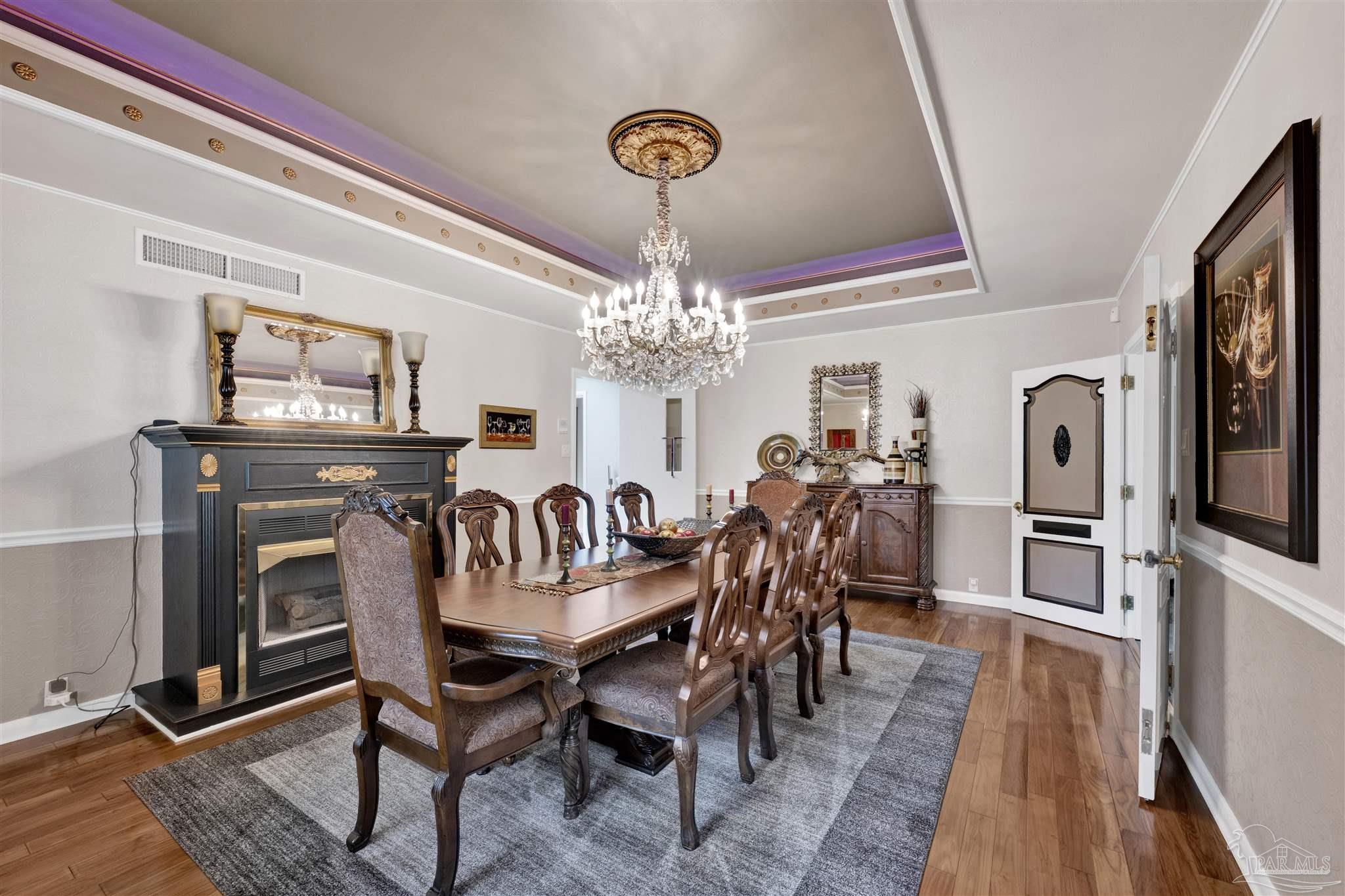 This formal dining room features a large wooden table surrounded by ornate, high-backed chairs, set beneath a grand crystal chandelier. A black fireplace with a decorative mantel and mirror serves as a focal point, while a matching wooden sideboard sits against the far wall. The room is defined by a tray ceiling with purple accent lighting and a large area rug over polished wood floors, creating an elegant and traditional atmosphere.