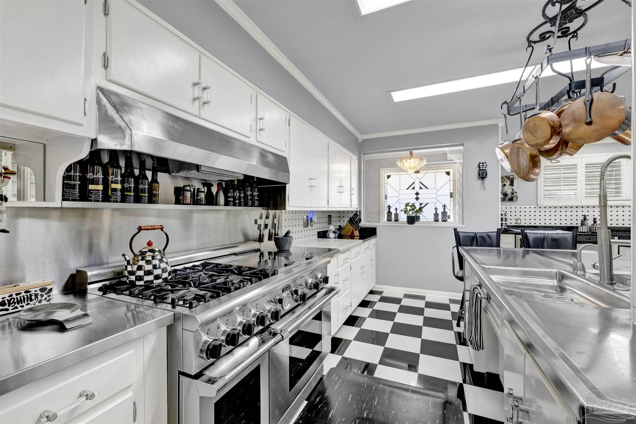 This kitchen features a striking black-and-white checkered floor and a professional-grade stainless steel range with a matching vent hood. The space is equipped with white cabinetry, stainless steel countertops, and a hanging rack for copper cookware, creating a functional and high-contrast aesthetic. A window with decorative leaded glass adds a unique architectural detail to the end of the galley-style layout.