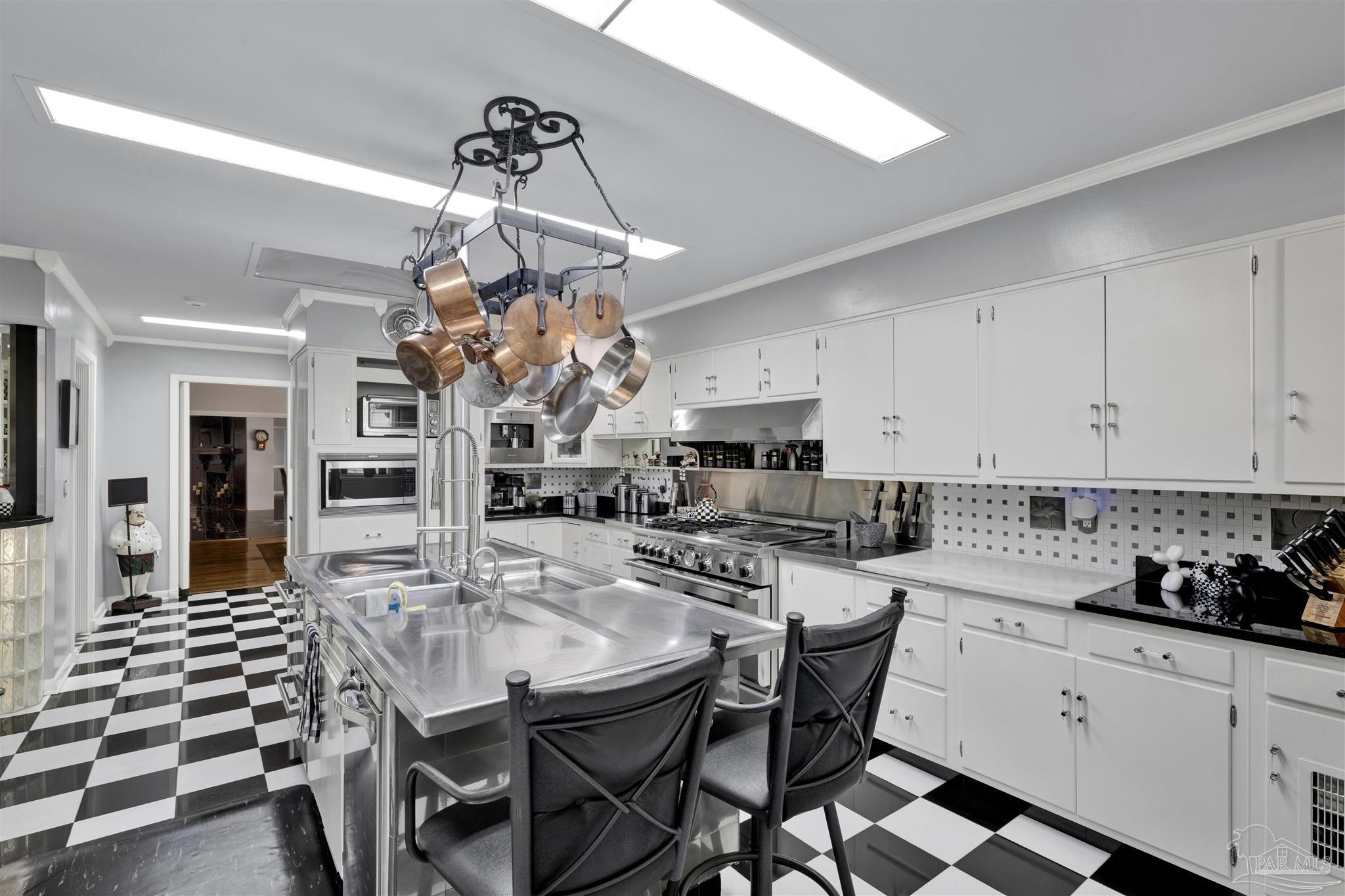 This kitchen features a striking black-and-white checkered floor and a large central stainless steel island with an integrated sink and bar seating. White cabinetry lines the walls, complemented by a professional-grade range and a hanging pot rack filled with copper cookware. The space is illuminated by long, rectangular recessed ceiling lights, creating a bright and functional culinary environment.