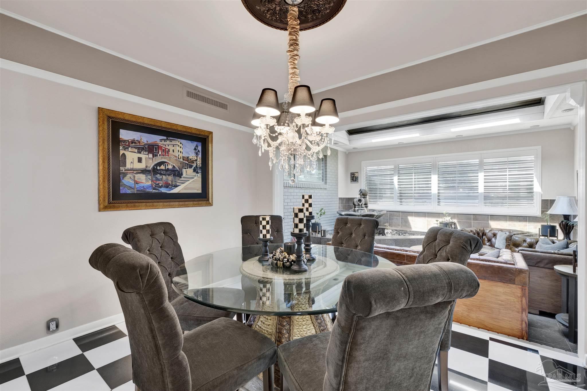 This elegant dining room features a round glass-top table surrounded by four tufted, charcoal-gray upholstered chairs, set against a striking black-and-white checkered floor. A crystal chandelier hangs from a decorative ceiling medallion, while a framed painting of a Venetian canal scene adds a touch of classic sophistication to the neutral-toned walls. The space opens into an adjacent living area, creating an inviting and cohesive flow between rooms.
