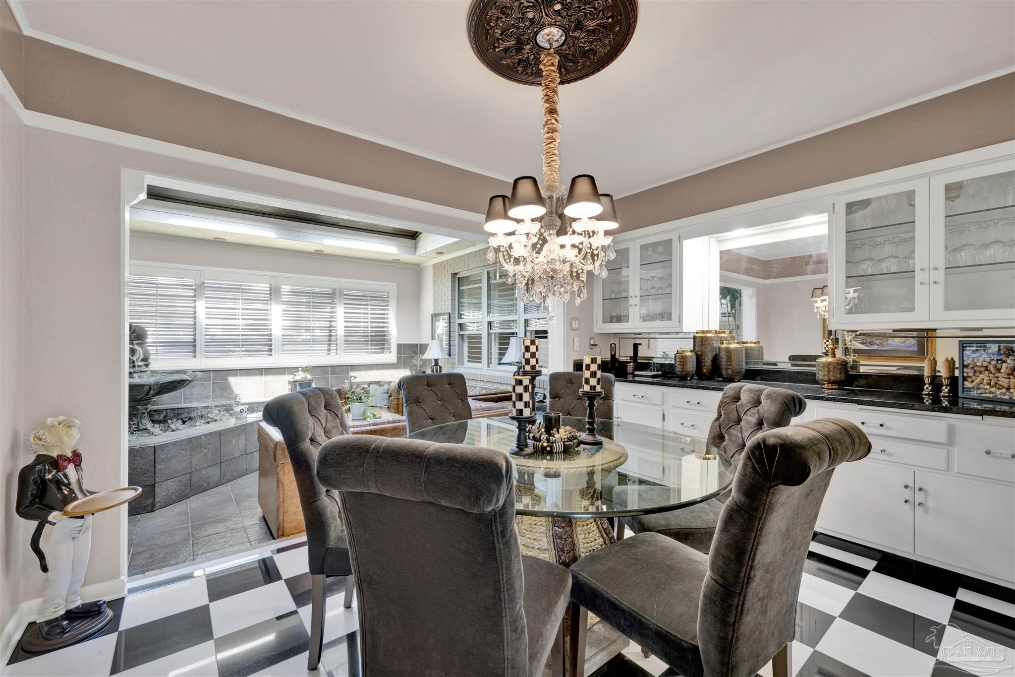 This elegant dining room features a glass-topped table surrounded by four tufted, dark gray upholstered chairs, set against a striking black-and-white checkered floor. A crystal chandelier hangs from a decorative ceiling medallion, while a butler-style statue stands to the left, adding a whimsical touch. The space opens into an adjacent sunroom area with a water feature, and white cabinetry with glass-front doors provides ample storage and display space.