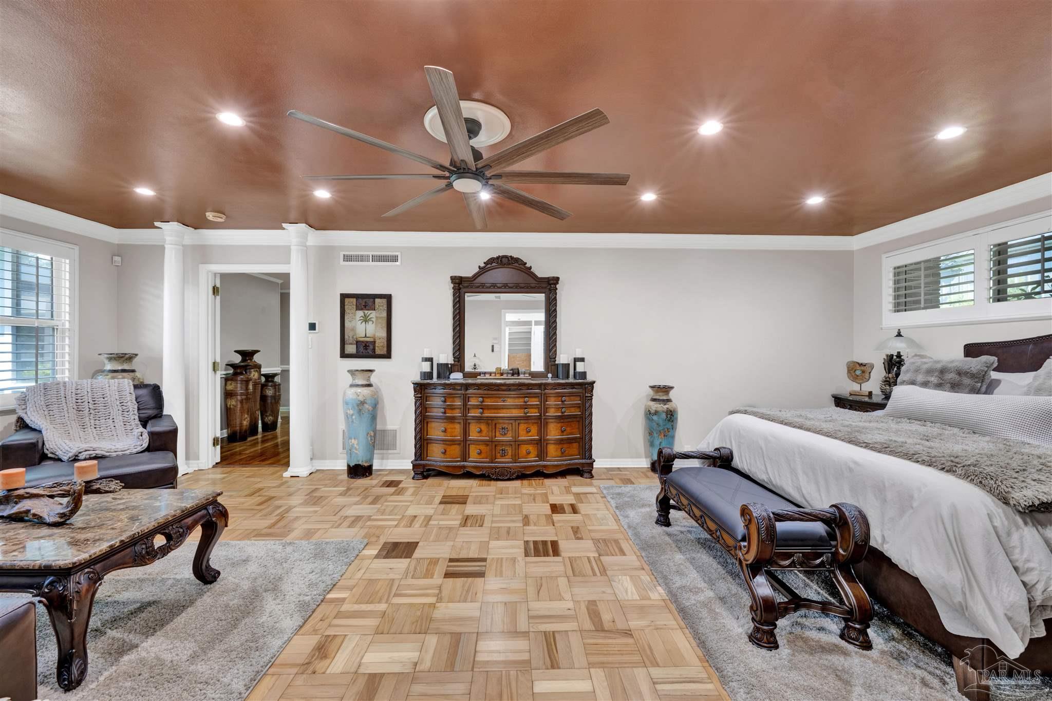 This spacious primary bedroom features a distinctive copper-toned ceiling with recessed lighting and a large ceiling fan. The room is anchored by a wooden dresser with an ornate mirror, flanked by decorative blue vases, and includes a large bed with a dark headboard and a bench at the foot. The flooring consists of a unique parquet wood pattern, complemented by area rugs and a seating area with a coffee table in the foreground.