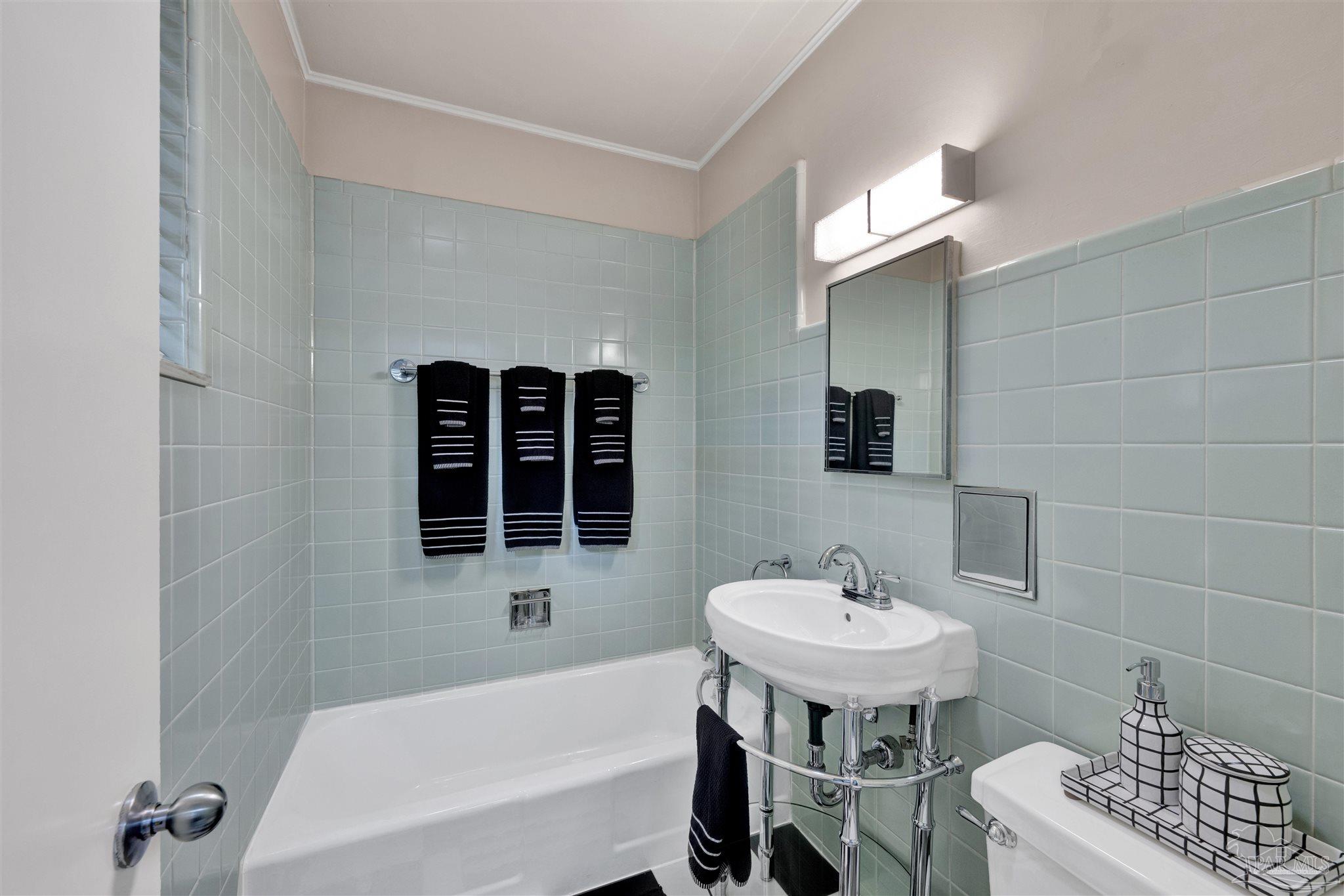 This vintage-inspired bathroom features light blue square wall tiles and a classic pedestal sink with chrome plumbing fixtures. A white bathtub is positioned against the tiled wall, complemented by a wall-mounted mirror and a rectangular light fixture above. The space is accented with black and white patterned towels and accessories, creating a clean and retro aesthetic.