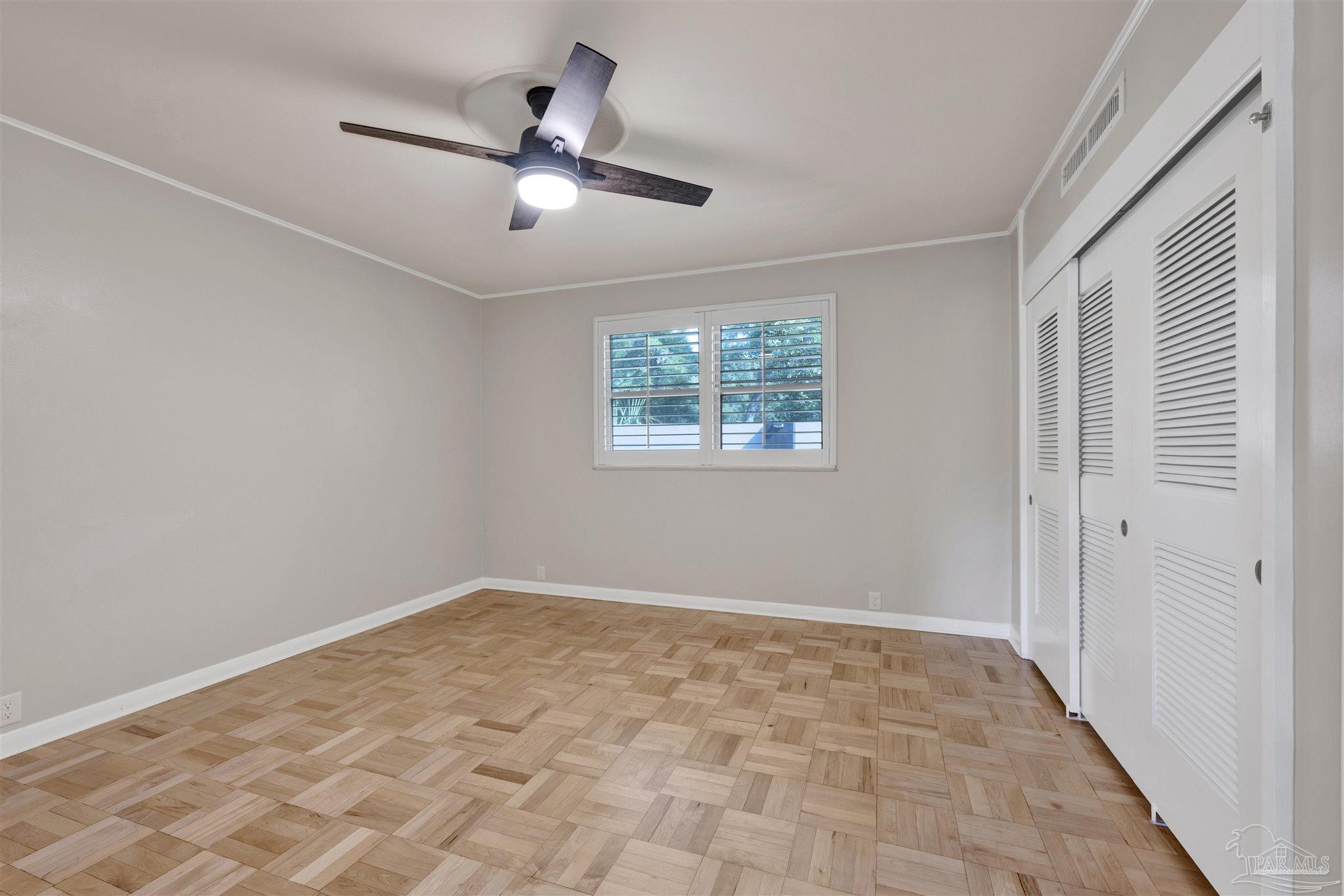This bright, neutral-toned bedroom features distinctive light wood parquet flooring and a central ceiling fan with integrated lighting. The room includes a set of white louvered closet doors on the right and a window with plantation-style shutters that allow natural light to filter in. The space is clean and minimalist, offering a versatile canvas for various interior design styles.