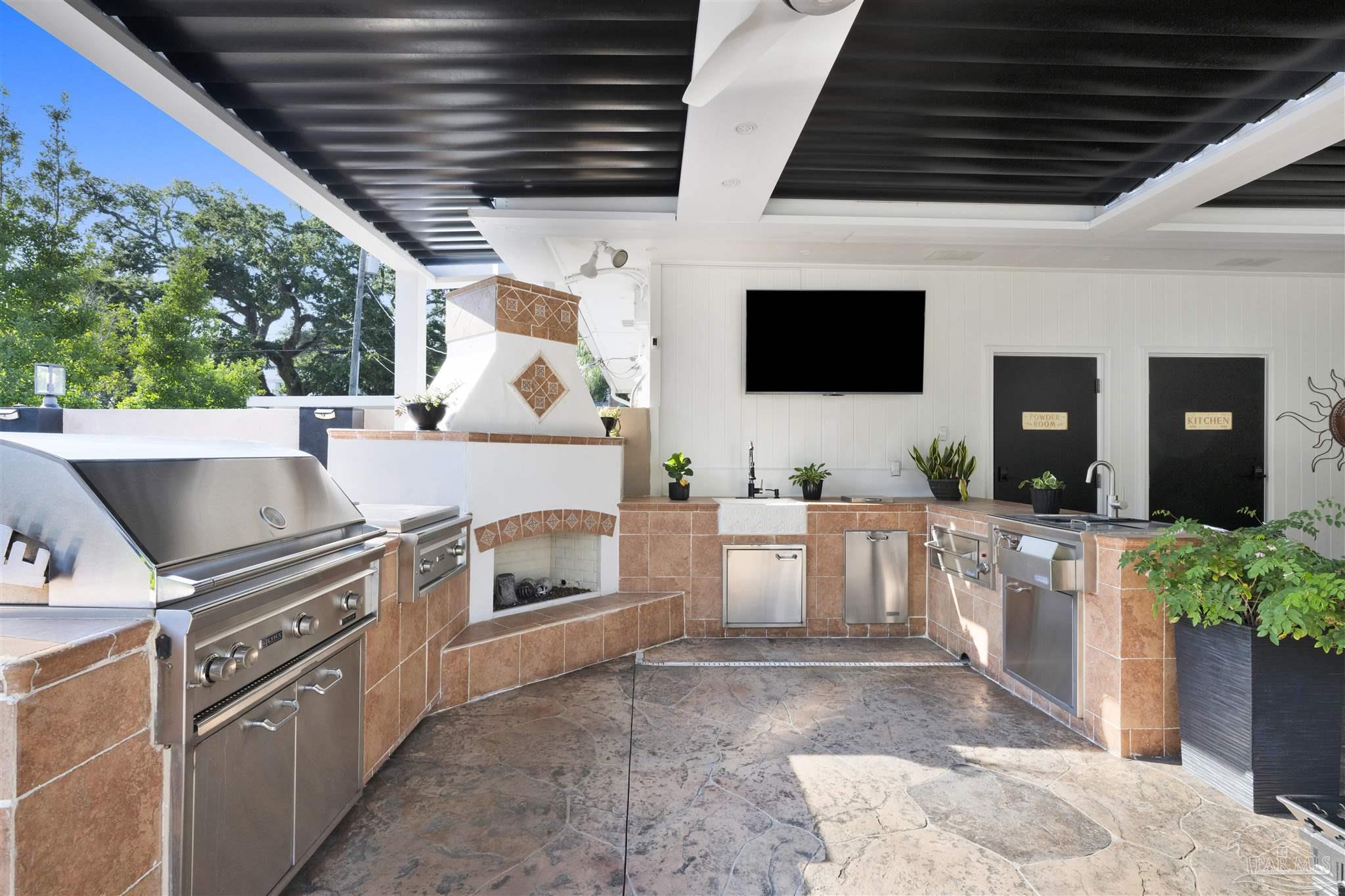 This expansive outdoor kitchen and living area features a covered patio with a dark slatted ceiling and a stone-tiled floor. The space is equipped with a large stainless steel grill, a built-in fireplace with decorative tile accents, and a sink area with stainless steel appliances. A wall-mounted television and two doors labeled 'Powder Room' and 'Kitchen' complete this well-appointed, functional entertainment space.