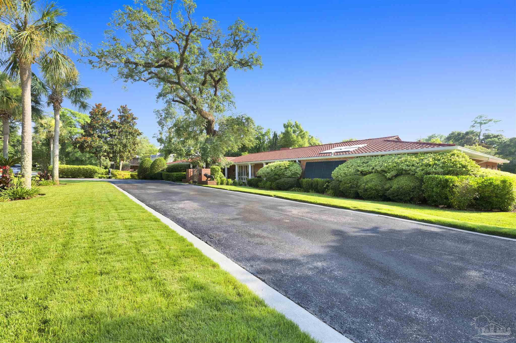 This image captures the front exterior of a sprawling, single-story residence featuring a classic red-tiled roof and a long, paved driveway leading toward the entrance. The property is surrounded by lush, manicured landscaping, including mature trees and dense, well-trimmed hedges that provide privacy. The perspective is from the driveway, emphasizing the home's wide footprint and inviting, sun-drenched curb appeal under a clear blue sky.