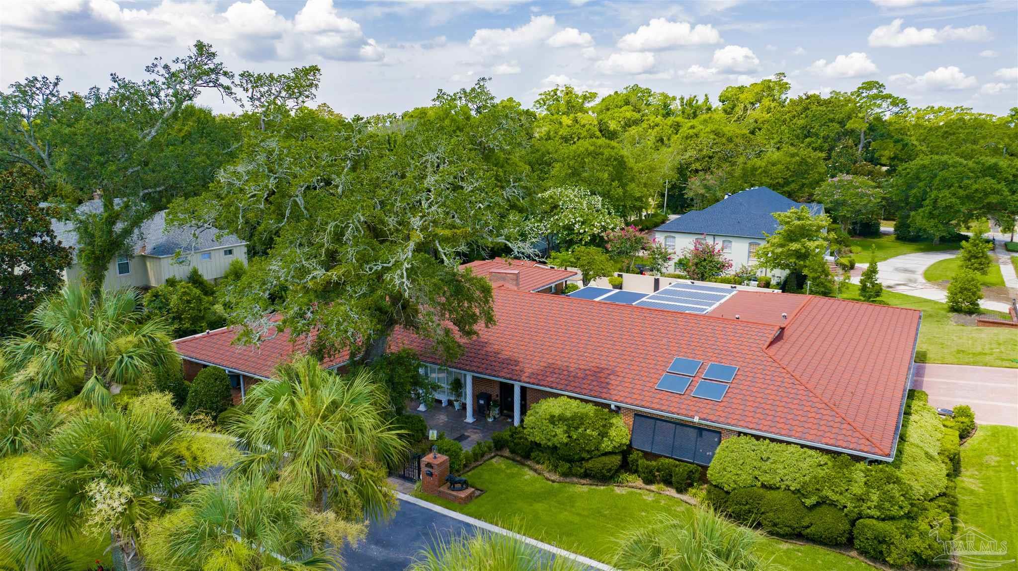 This aerial view captures a sprawling single-story residence with a distinctive red tile roof, set amidst a lush, tree-filled landscape. The home features solar panels on the roof, a covered front entryway, and a well-manicured lawn, all framed by mature oak trees and dense greenery. The perspective provides a clear overview of the property's layout and its integration into the surrounding suburban environment.