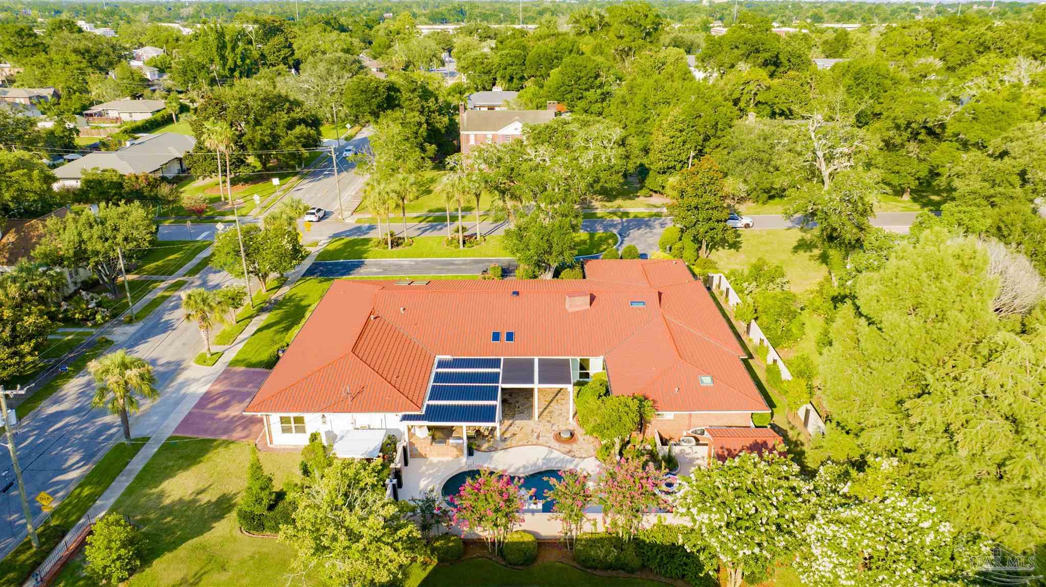 This high-angle aerial view captures a sprawling single-story residence with a prominent red metal roof, featuring integrated solar panels and skylights. The property includes a private swimming pool set within a paved patio area, surrounded by lush green landscaping and mature trees. The perspective highlights the home's layout and its integration into a quiet, tree-lined suburban neighborhood.