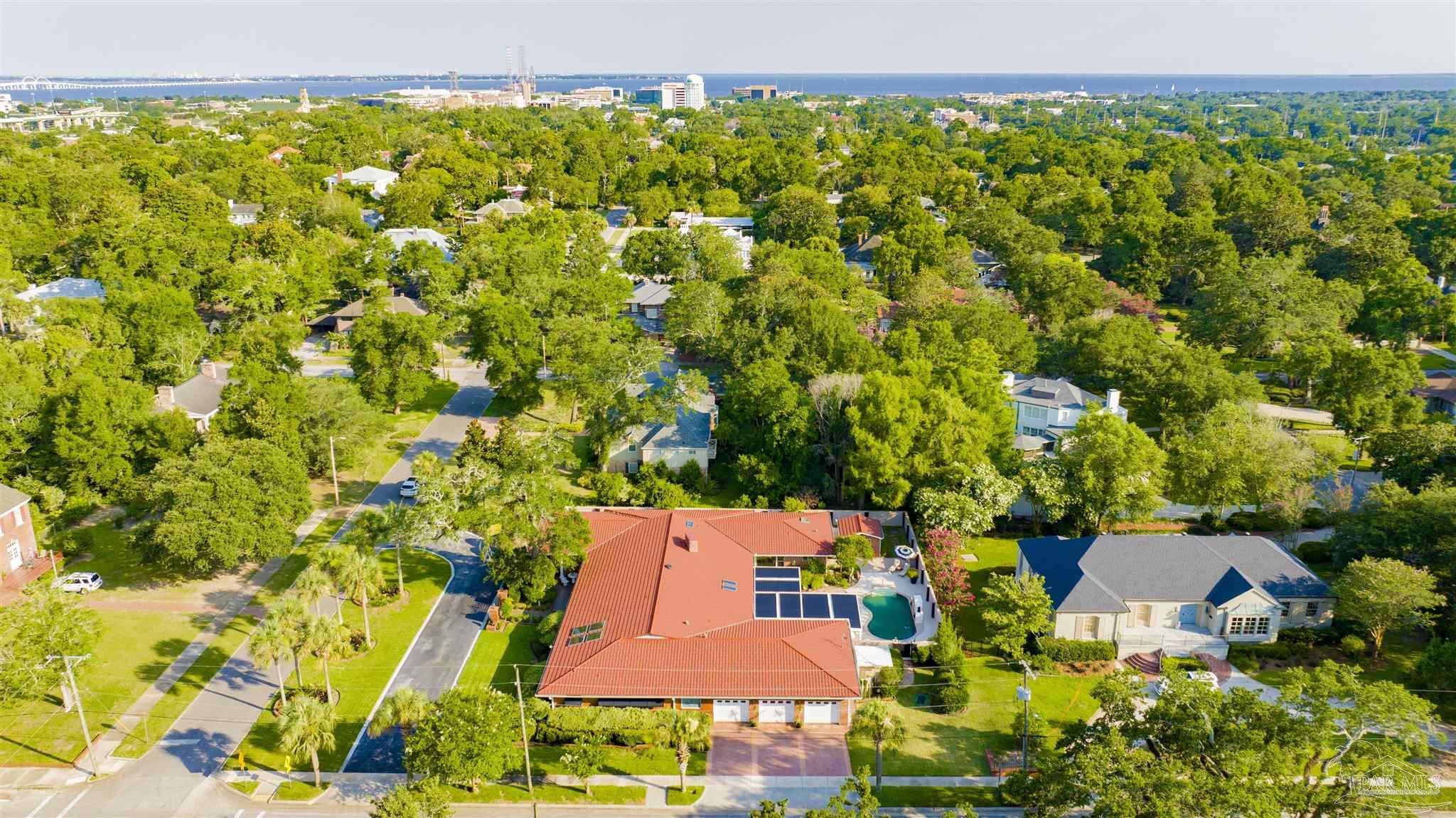 This high-angle aerial view captures a sprawling residential property featuring a large home with a prominent red tile roof and an attached three-car garage. The backyard includes a private swimming pool and patio area, all surrounded by lush, mature trees and well-manicured landscaping. The perspective provides a clear look at the neighborhood layout, with the distant coastline and water visible on the horizon.