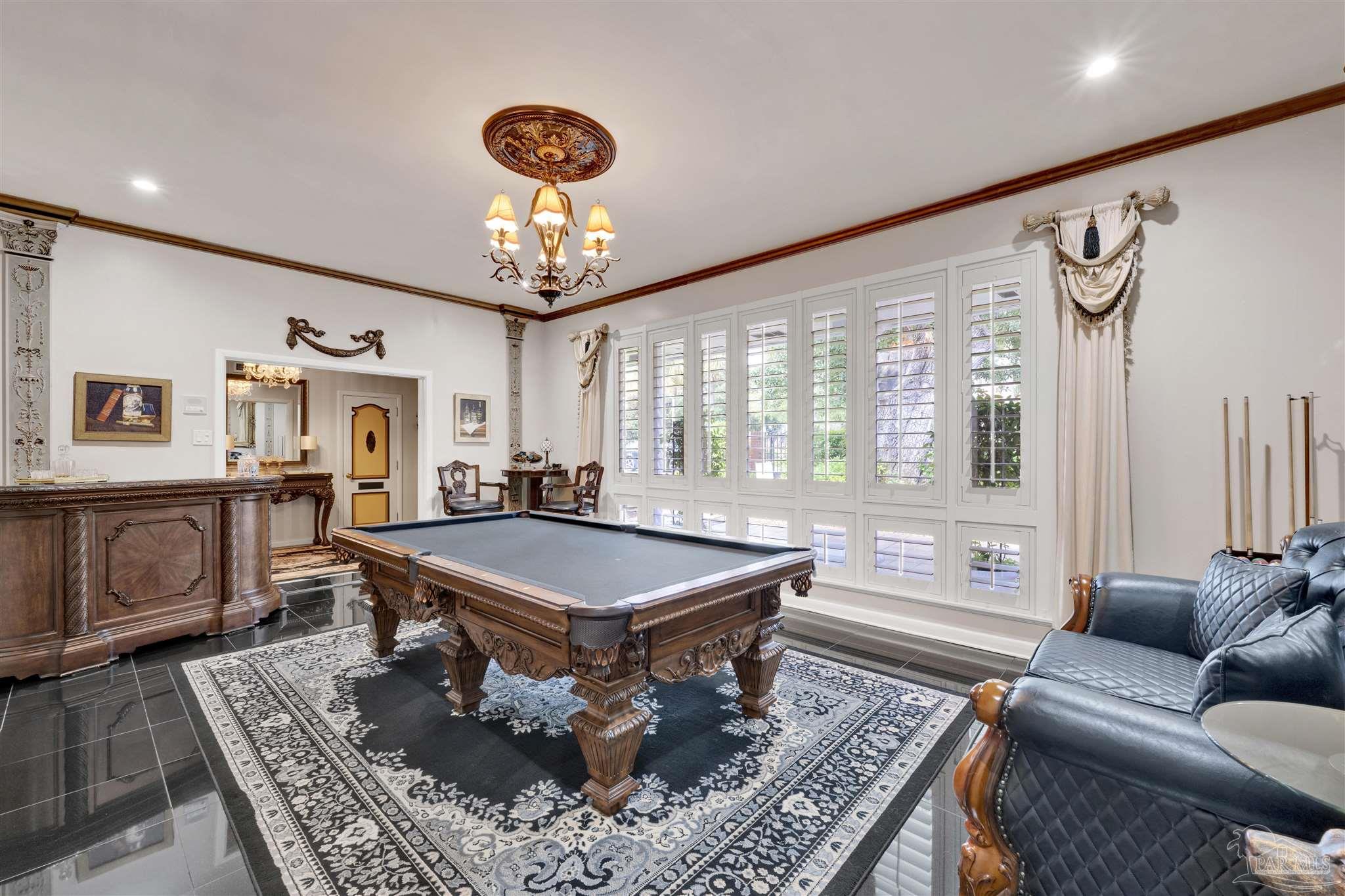 This elegant game room features a central pool table set atop a large, ornate patterned rug on a dark, polished tile floor. The space is framed by tall, white-shuttered windows and includes a dark wood bar area, a tufted leather sofa, and a decorative chandelier hanging from a detailed ceiling medallion. The room exudes a formal, classic aesthetic with rich wood tones and sophisticated architectural accents.