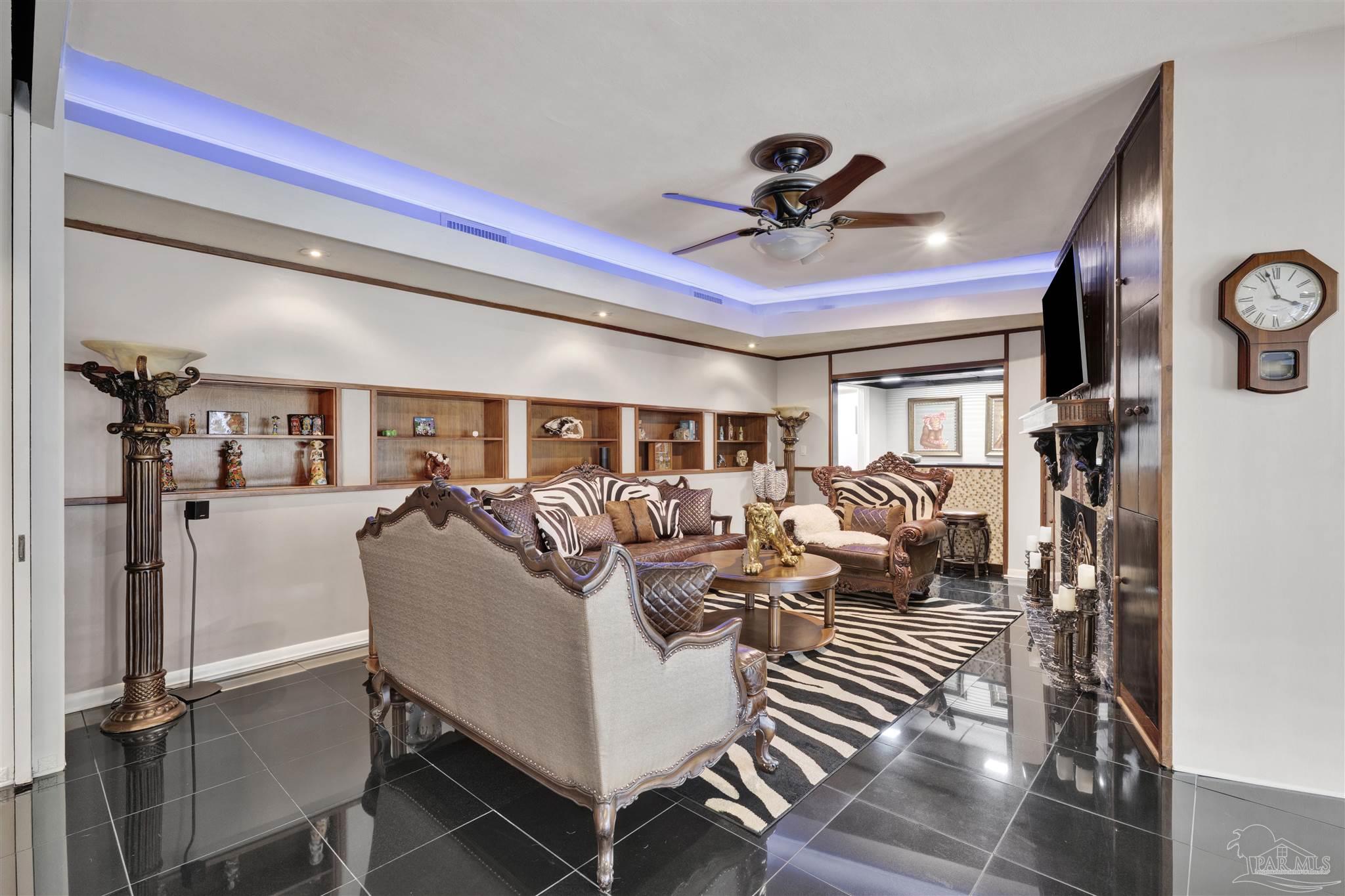 This opulent living room features a bold, eclectic design with high-gloss black tile flooring and a striking zebra-print area rug. The space is anchored by ornate, dark-wood furniture with intricate carvings and is illuminated by recessed lighting with a blue LED accent cove. A built-in shelving unit spans the wall, while a wall-mounted clock and a decorative pedestal lamp add to the room's dramatic, formal aesthetic.
