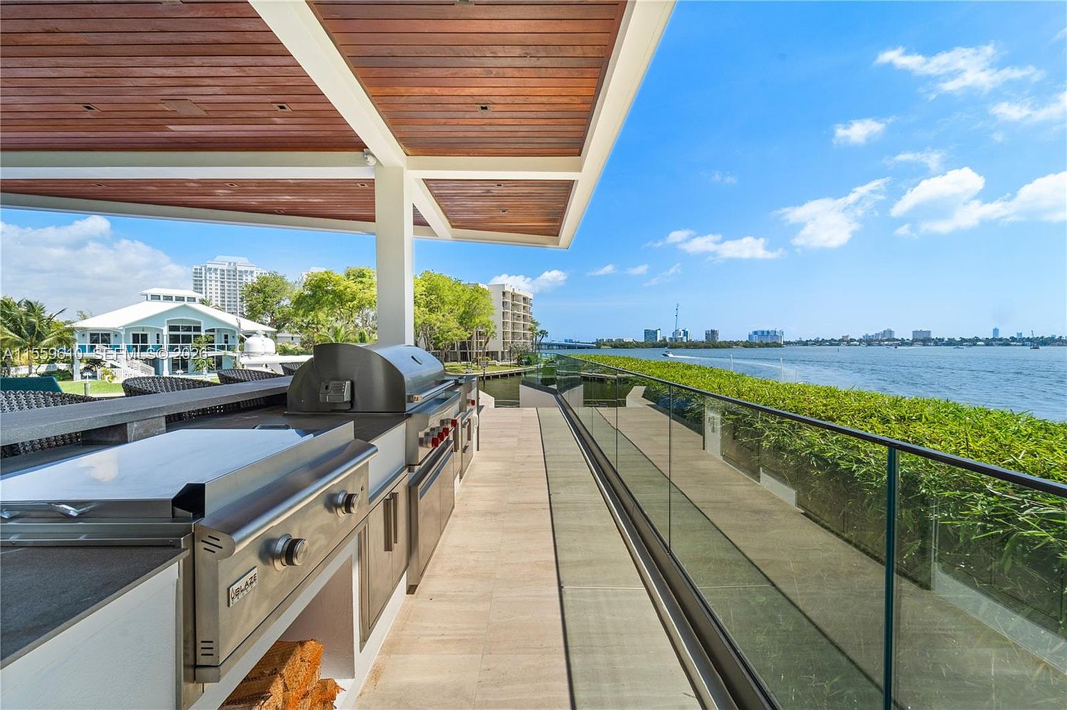 This image showcases a luxurious outdoor kitchen and patio area with a stunning waterfront view. The space features high-end stainless steel appliances, a sleek countertop, and a modern pergola overhead with wooden slats. A glass railing provides an unobstructed view of the water and cityscape beyond, creating an inviting and sophisticated outdoor living space.