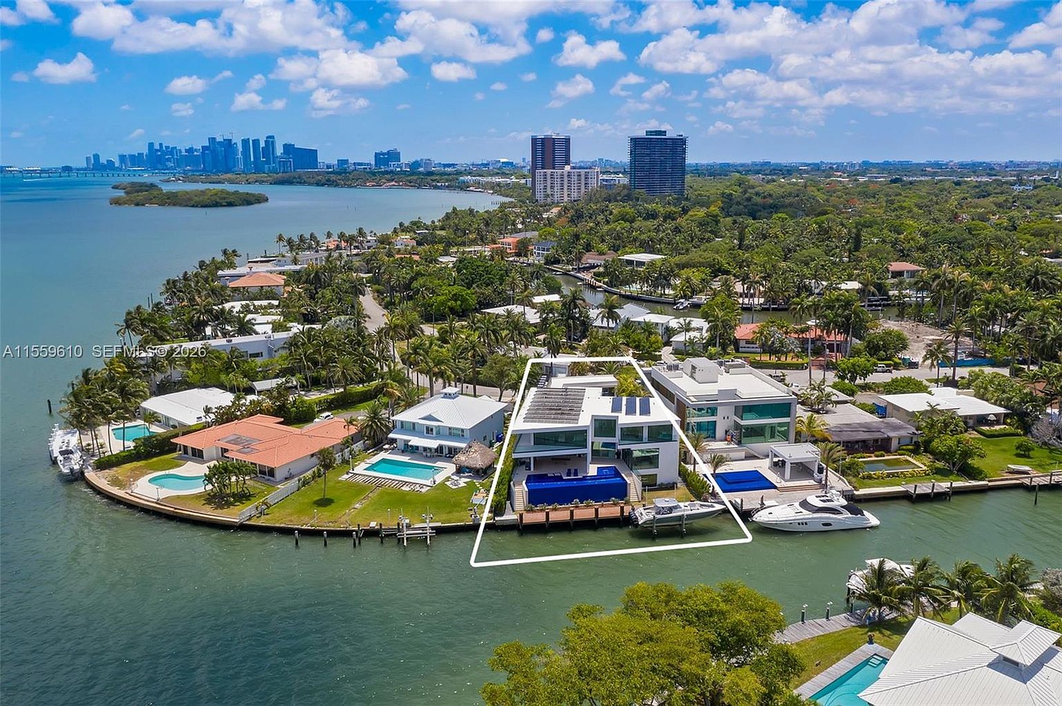 This aerial shot showcases a luxurious waterfront property with a modern architectural design. The house features a flat roof with solar panels, large windows, and a private dock with boats. The surrounding landscape includes lush greenery, a canal, and a view of the city skyline in the distance, creating an impression of exclusivity and high-end living.