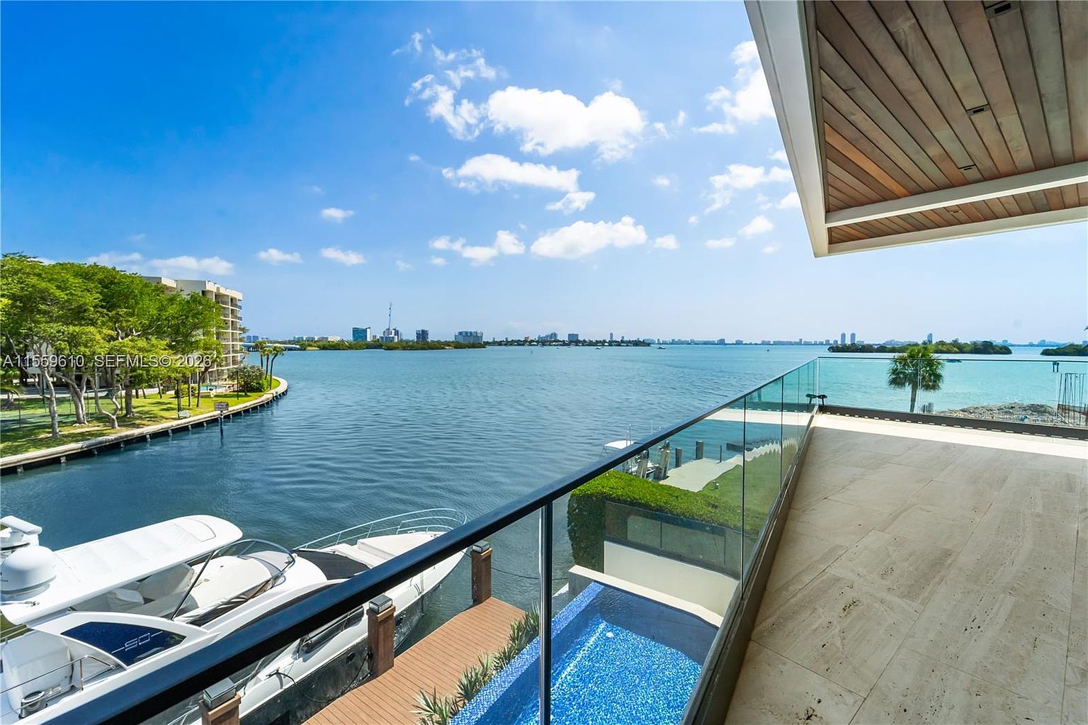 This image showcases a luxurious patio or balcony with stunning waterfront views. The space features a glass railing, a small pool with blue mosaic tiles, and a wooden deck area. Several boats are docked nearby, adding to the upscale ambiance. The view extends to a cityscape in the distance under a partly cloudy sky.