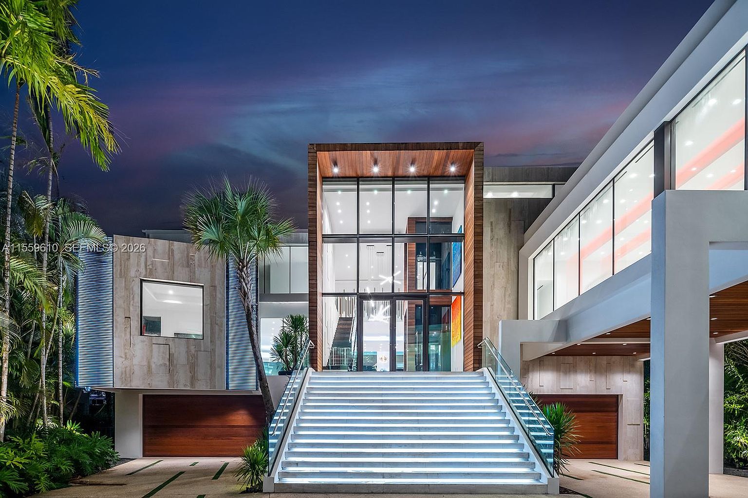 This is a striking front view of a modern luxury home at dusk. The property features a grand entrance with illuminated stairs leading to a multi-story glass facade framed by wood and concrete elements. Palm trees and manicured landscaping add to the upscale curb appeal, while the overall design exudes sophistication and contemporary elegance.