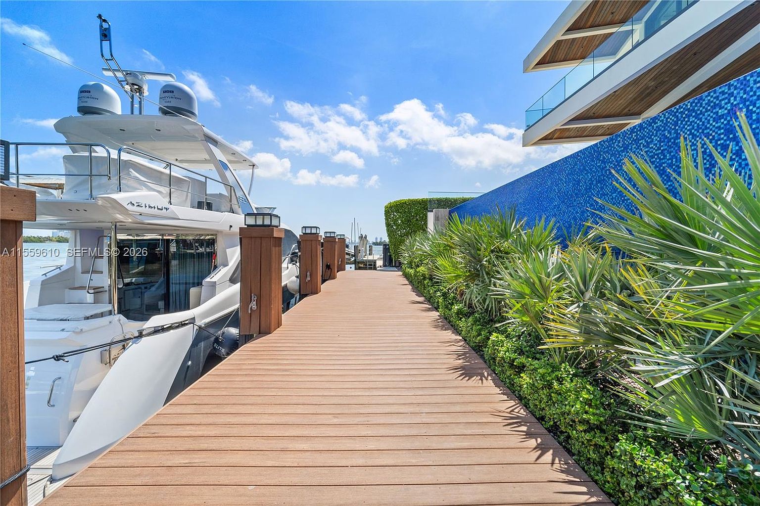 This image showcases a luxurious waterfront property featuring a private dock with a large yacht moored alongside. The dock is constructed of rich, warm-toned wood and is lined with manicured greenery and a striking blue tiled wall. The scene is bathed in sunlight, creating a sense of opulence and tranquility, perfect for a high-end real estate listing.