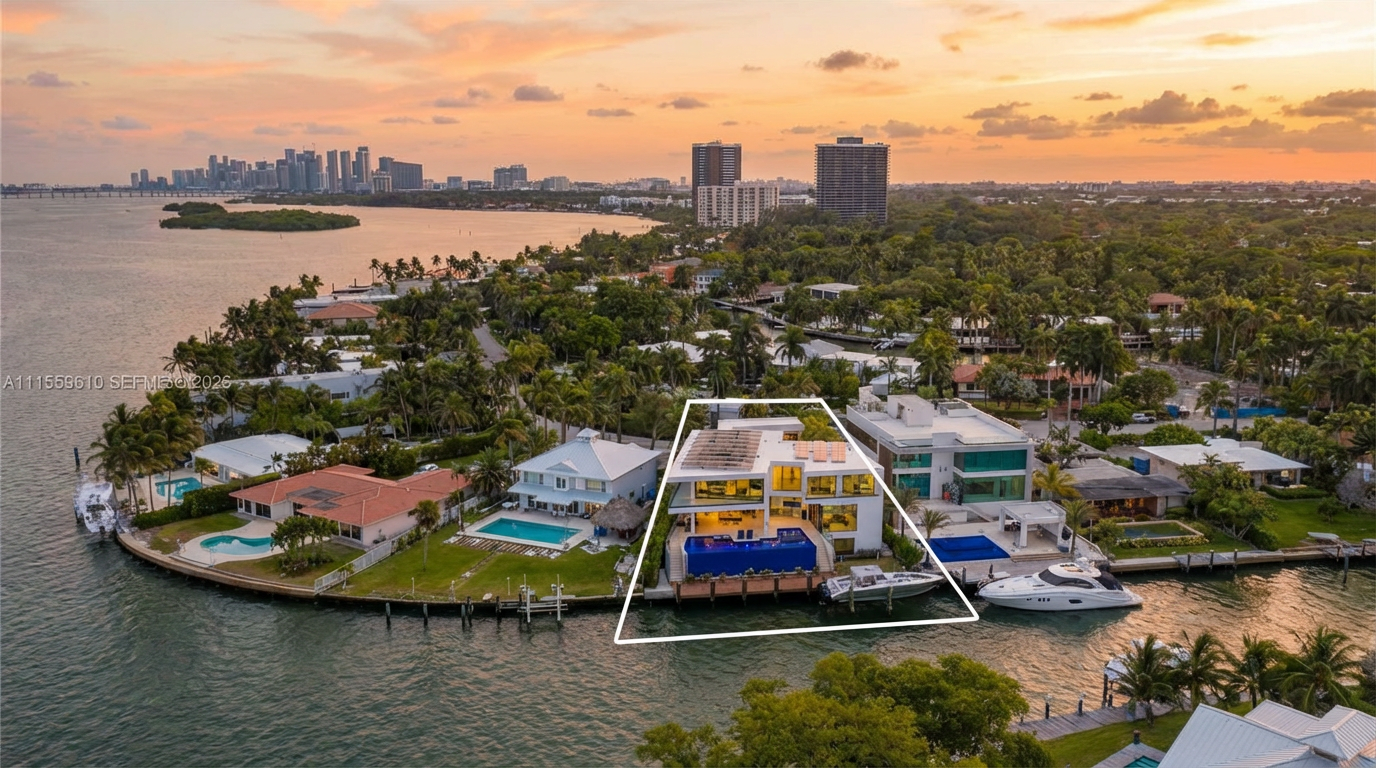 This stunning aerial view highlights a contemporary waterfront estate designed with modern architectural lines and expansive glass windows. The property features a prominent infinity-edge pool overlooking the bay, complemented by a private dock and a prime location in a lush, tropical residential enclave with iconic city views in the backdrop.