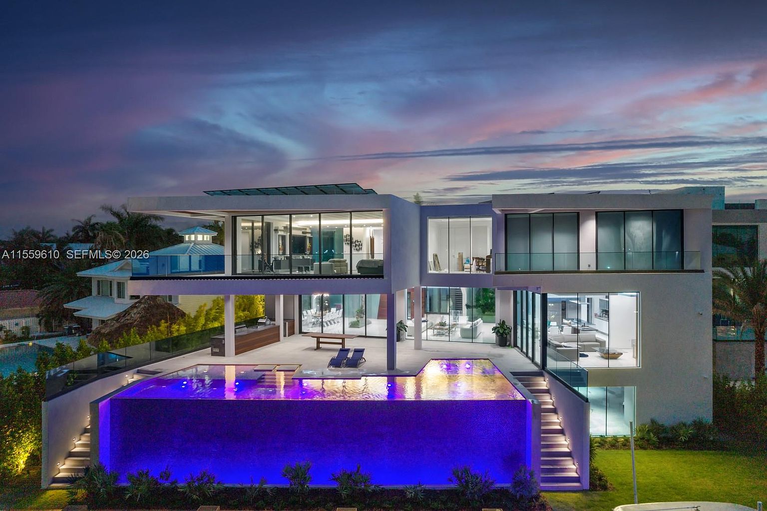 This is a rear exterior view of a modern, multi-story home featuring expansive glass windows and a striking infinity pool illuminated with blue lighting. The architecture emphasizes clean lines and open spaces, with outdoor living areas seamlessly integrated into the design. The property is well-landscaped, enhancing its luxurious and contemporary appeal.
