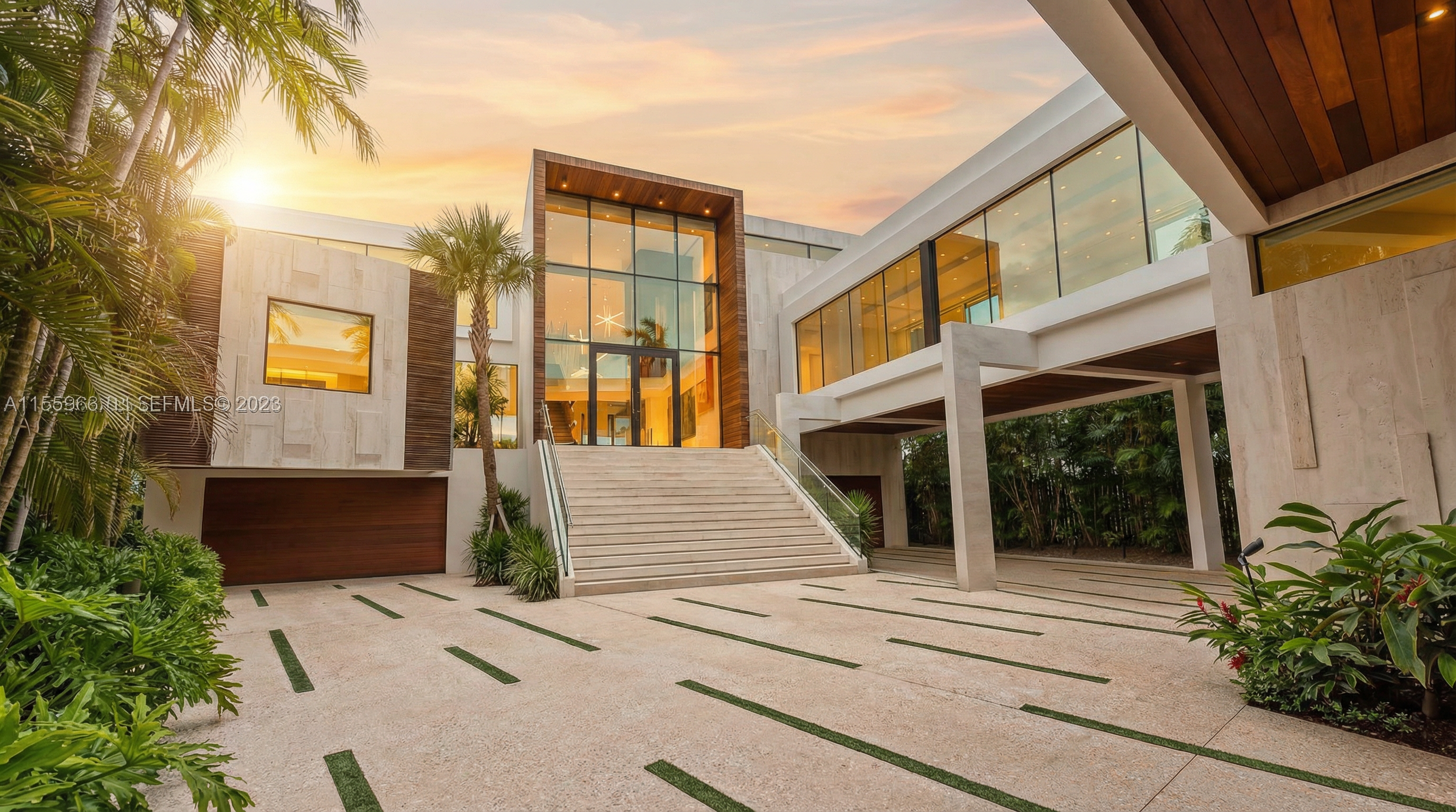 This stunning contemporary estate features a grand, elevated entryway with wide stone stairs and a towering glass facade. The residence combines sleek, modern architectural lines with natural elements like wood paneling and stone cladding, complemented by lush tropical landscaping and an inviting driveway. The sunset lighting enhances the luxurious and sophisticated curb appeal of this high-end property.