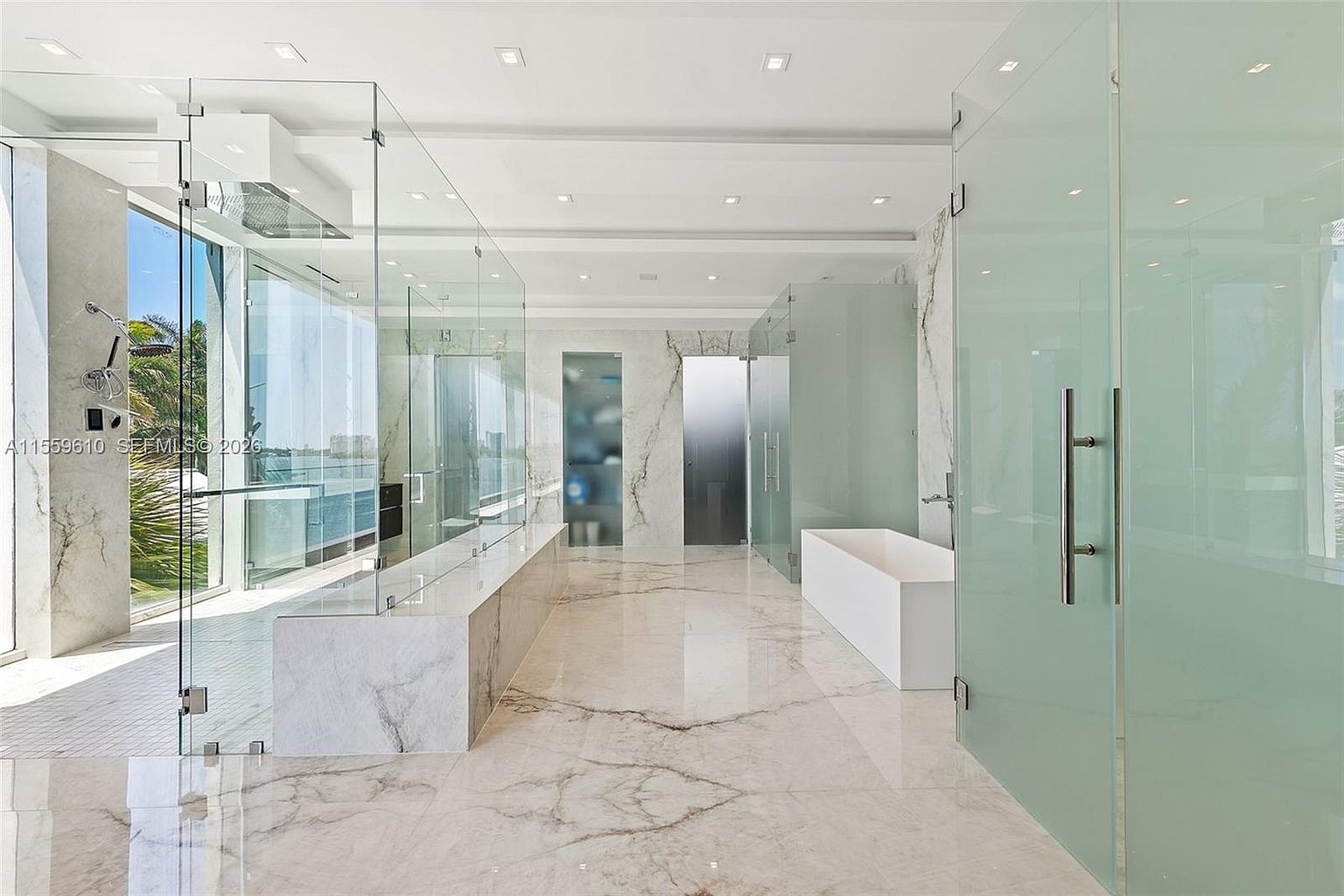 This is a luxurious primary bathroom featuring marble flooring and walls, glass-enclosed shower areas, and a modern freestanding bathtub. The design emphasizes clean lines and a spacious layout, creating a spa-like atmosphere. The perspective is from the doorway, showcasing the length of the bathroom and the various glass enclosures.