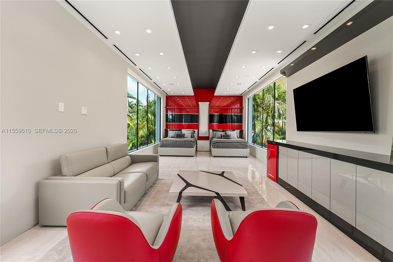 This is a modern bedroom featuring two beds with gray bedding, separated by a central red accent wall. The room has large windows offering views of lush greenery, and a sleek entertainment unit with a large television. The overall design is contemporary with a focus on clean lines and a bold color palette, creating a luxurious and inviting space.