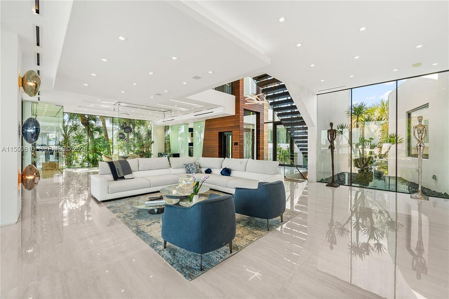 This is a spacious and modern living room featuring a large white sectional sofa, two blue armchairs, and a glass coffee table on a patterned rug. The room is bright with natural light from large windows overlooking lush greenery, and the polished tile floors reflect the light, enhancing the airy feel. A striking staircase and unique art pieces add to the room's sophisticated ambiance.