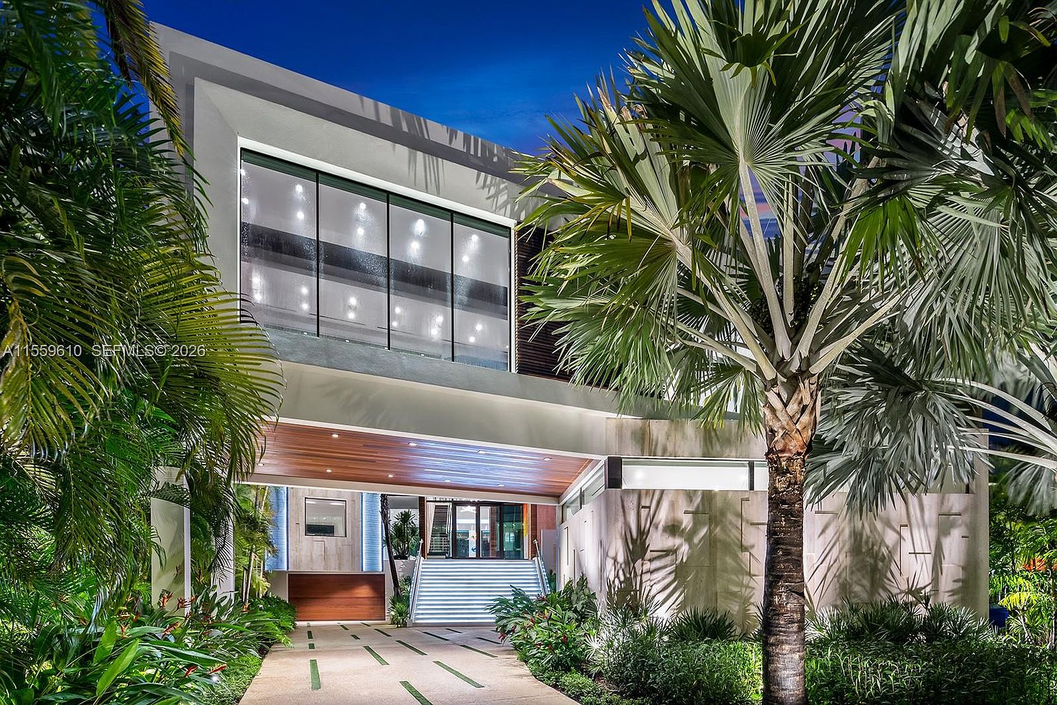 This is a front view of a modern, luxurious home. The architecture features clean lines, a large window, and a covered entryway with wooden accents. The landscaping is lush with tropical plants, creating an inviting and sophisticated curb appeal.