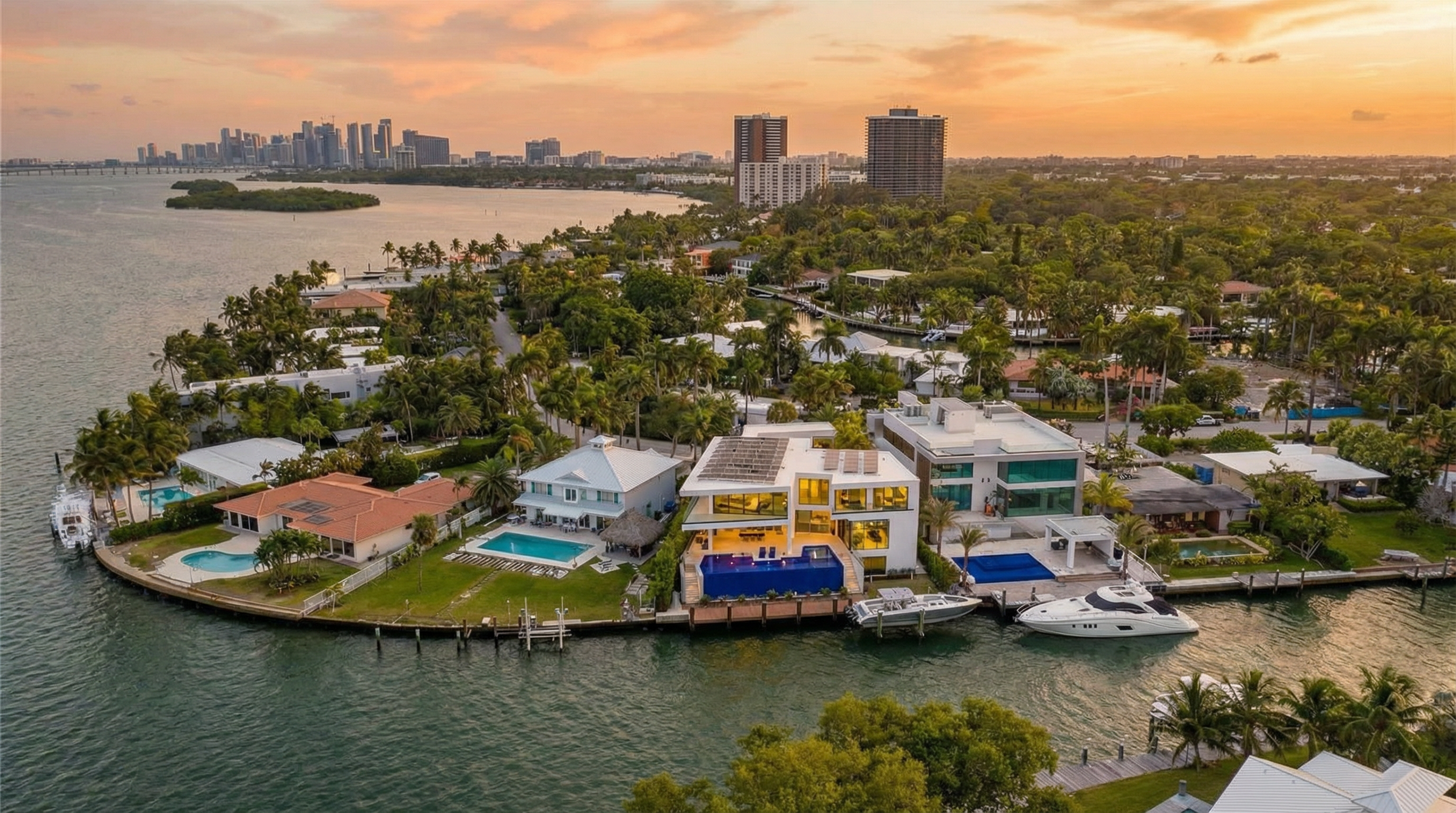 This stunning aerial view highlights a collection of luxury waterfront properties nestled along a winding canal in a tropical setting. The scene features modern and traditional architectural styles, private docks with yachts, and expansive swimming pools, all framed by lush palm trees and a sunset sky over the downtown skyline. The composition emphasizes the high-end, affluent lifestyle and desirable water access of this residential enclave.